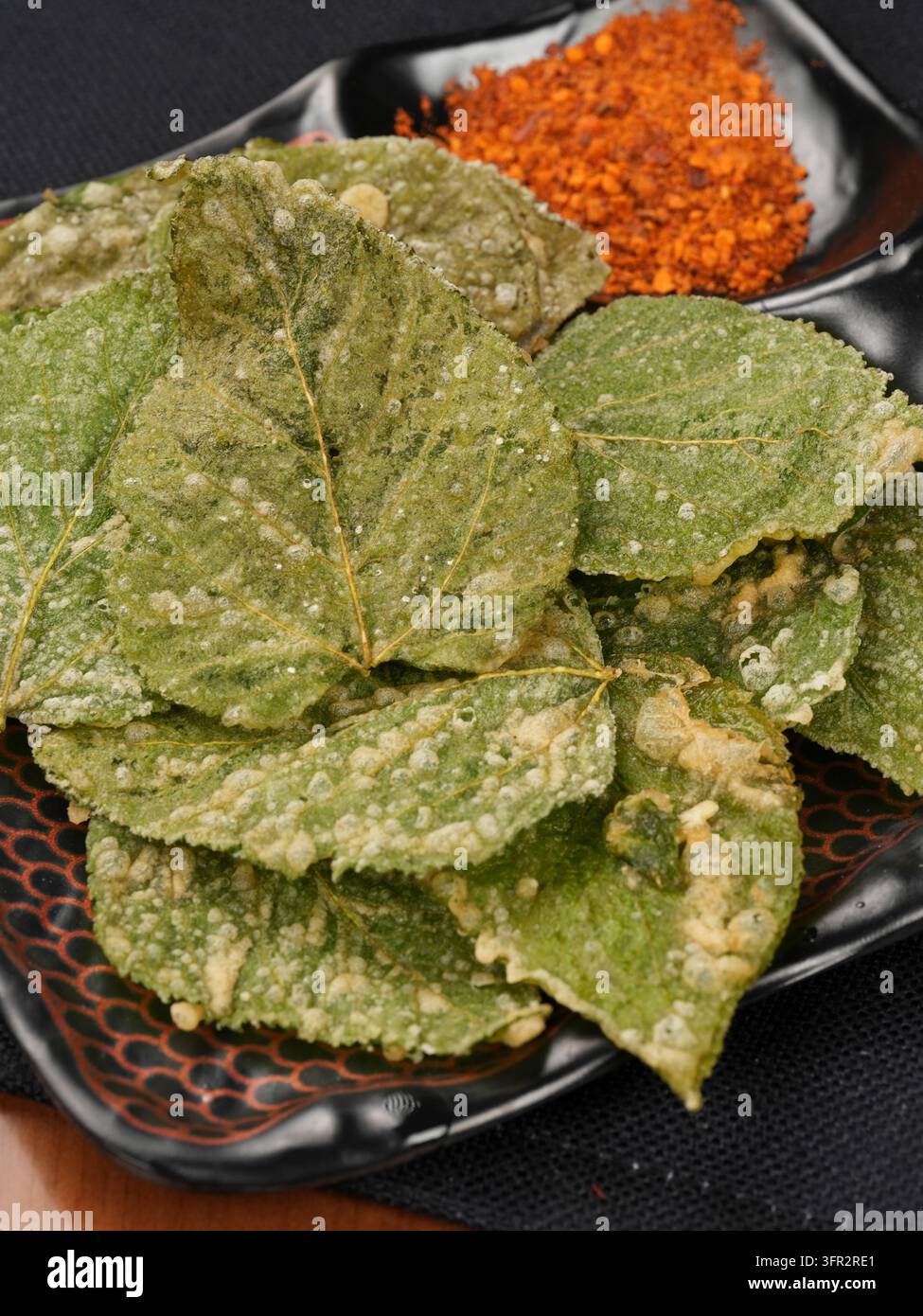 Nahaufnahme gebratener Maulbeerblätter auf schwarzem Teller mit zwei Fächern und Chilipulver Stockfoto