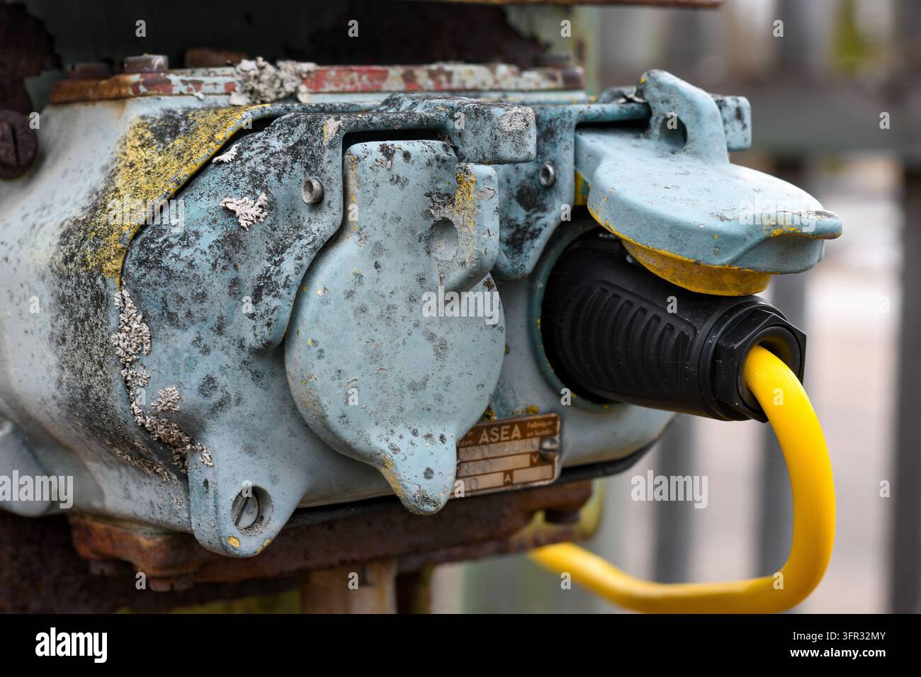 Nahaufnahme eines alten, verwitterten ASEA-Industriestromsockets mit einem gelben Stromkabel, das abgenutzte Farbe, Korrosion und Metallbeschläge zeigt. Stockfoto