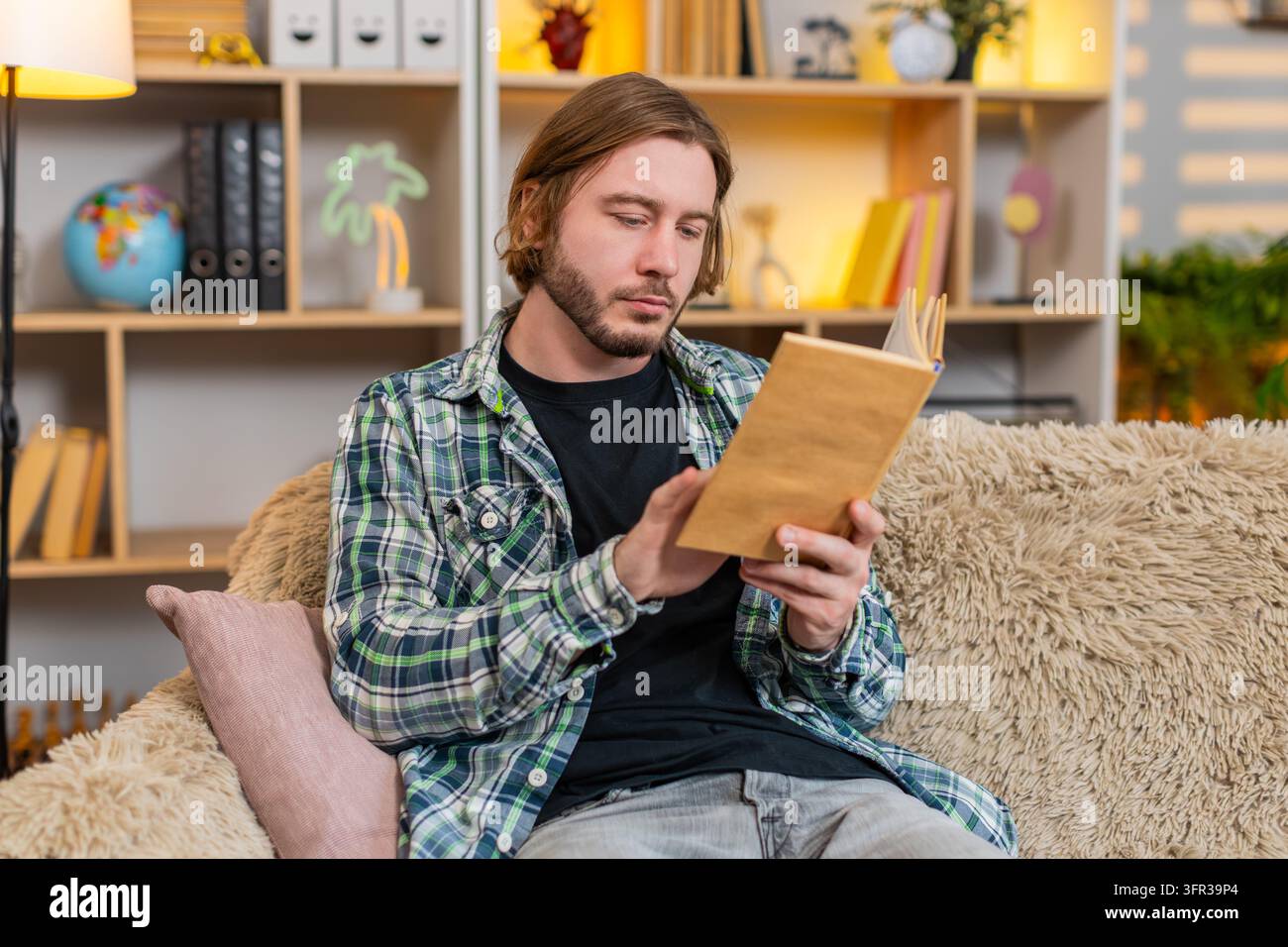 Junger Mann auf dem Heimsofa liest ein Buch mit warmem Lächeln und genießt ruhige Freizeit nach dem Arbeitsstress zu Hause. Erwachsener Mann auf dem Heimsofa findet Trost in einer inspirierenden Geschichte und ruhiger Ruhe im Haus während der Abendpause. Stockfoto