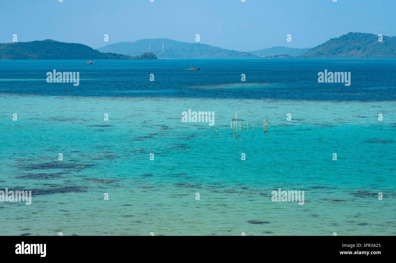 Klares türkisfarbenes tropisches Meer mit flachem Korallenwasser, entfernten grünen Inseln, kleinen Booten und einem ruhigen blauen Himmel. Stockfoto