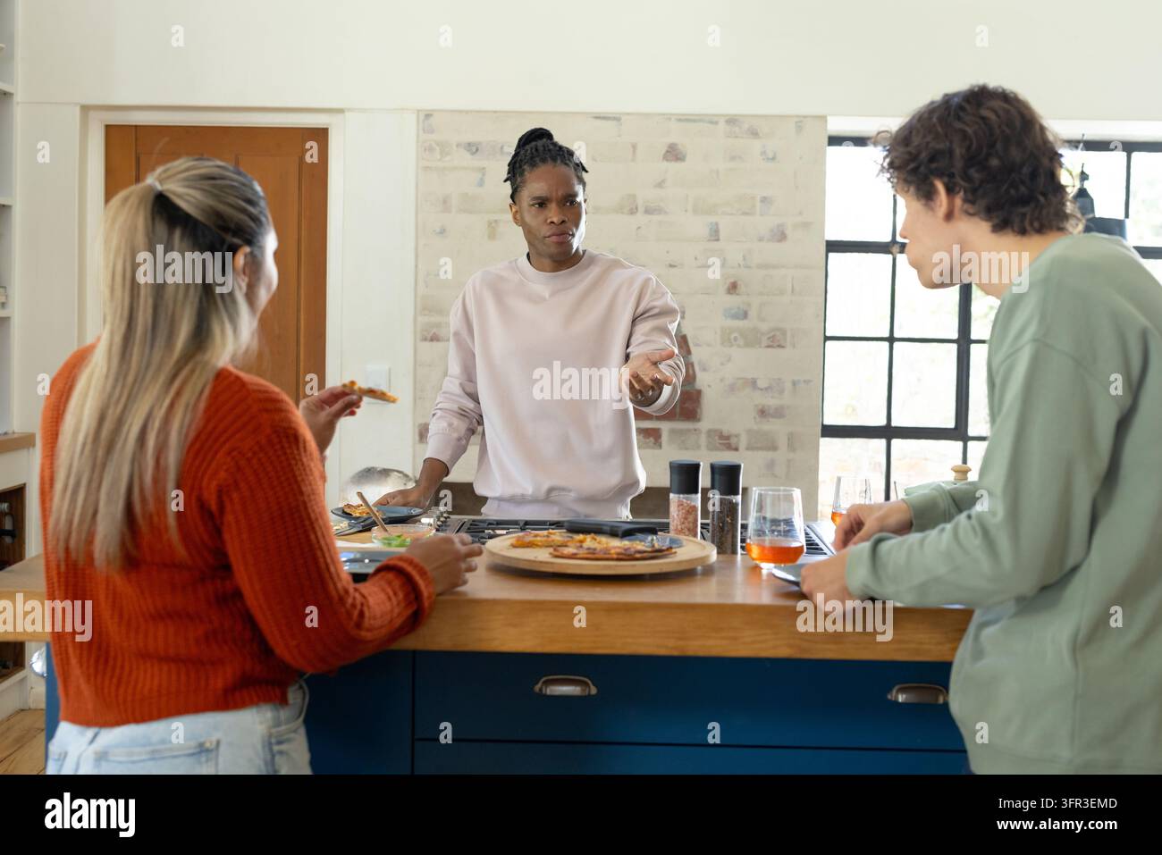 Verschiedene Freunde stehen um die Kücheninsel herum und kochen Pizza auf dem Grill mit Pfanne und Tablet in der Nähe Stockfoto