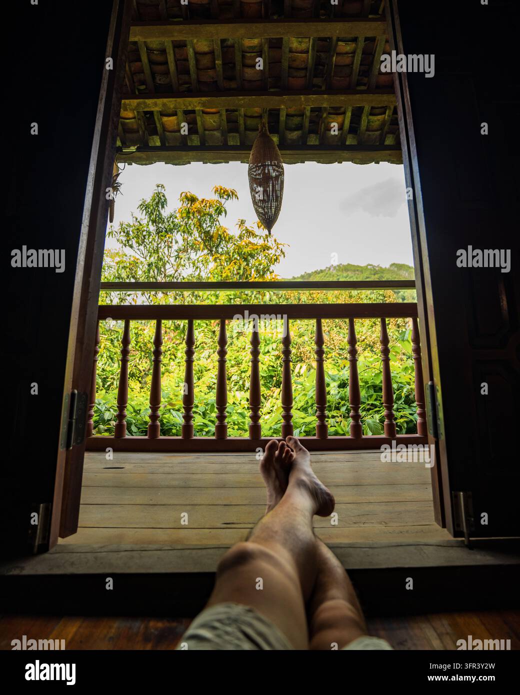 Ansichtspunkt von entspannten Beinen auf Holzboden eines Balkons, die durch offene Türen in den Garten schauen, Dong Lam, Huu Lung, Lang Son, Vietnam Stockfoto