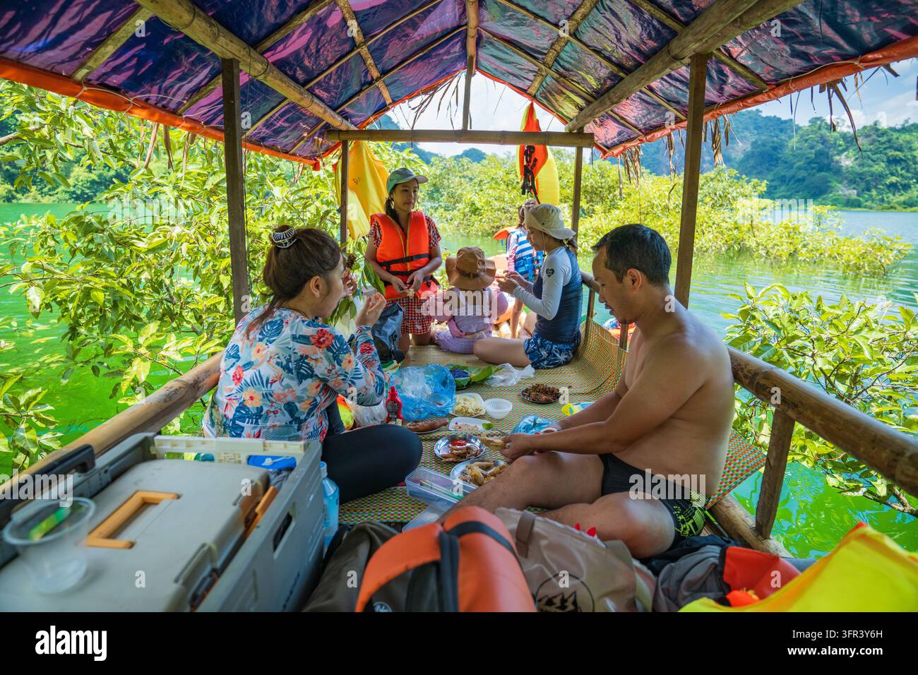Familie, die unter dem Baldachin eines Bambusfloßes auf einem grünen See ein Picknickessen einnimmt, mit Schwimmwesten und Kühlbox, Dong Lam, Huu Lung, Lang Son, Vietnam Stockfoto
