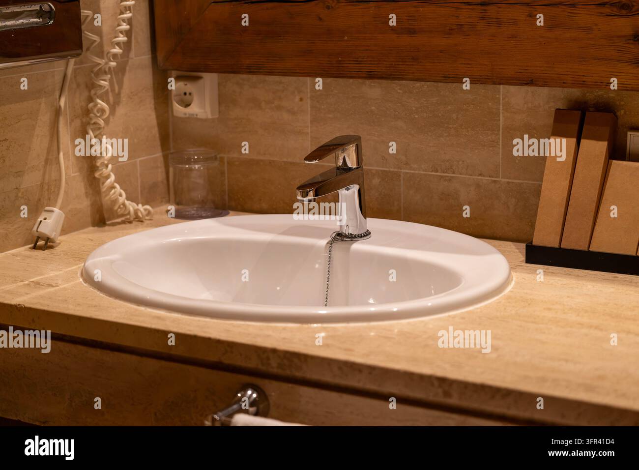 Weißes Keramik-Waschbecken mit einem Chrom-Wasserhahn, eingebaut in eine beige Stein-Arbeitsplatte, mit einem wandmontierten Telefon und einem Trinkglas im Hintergrund Stockfoto