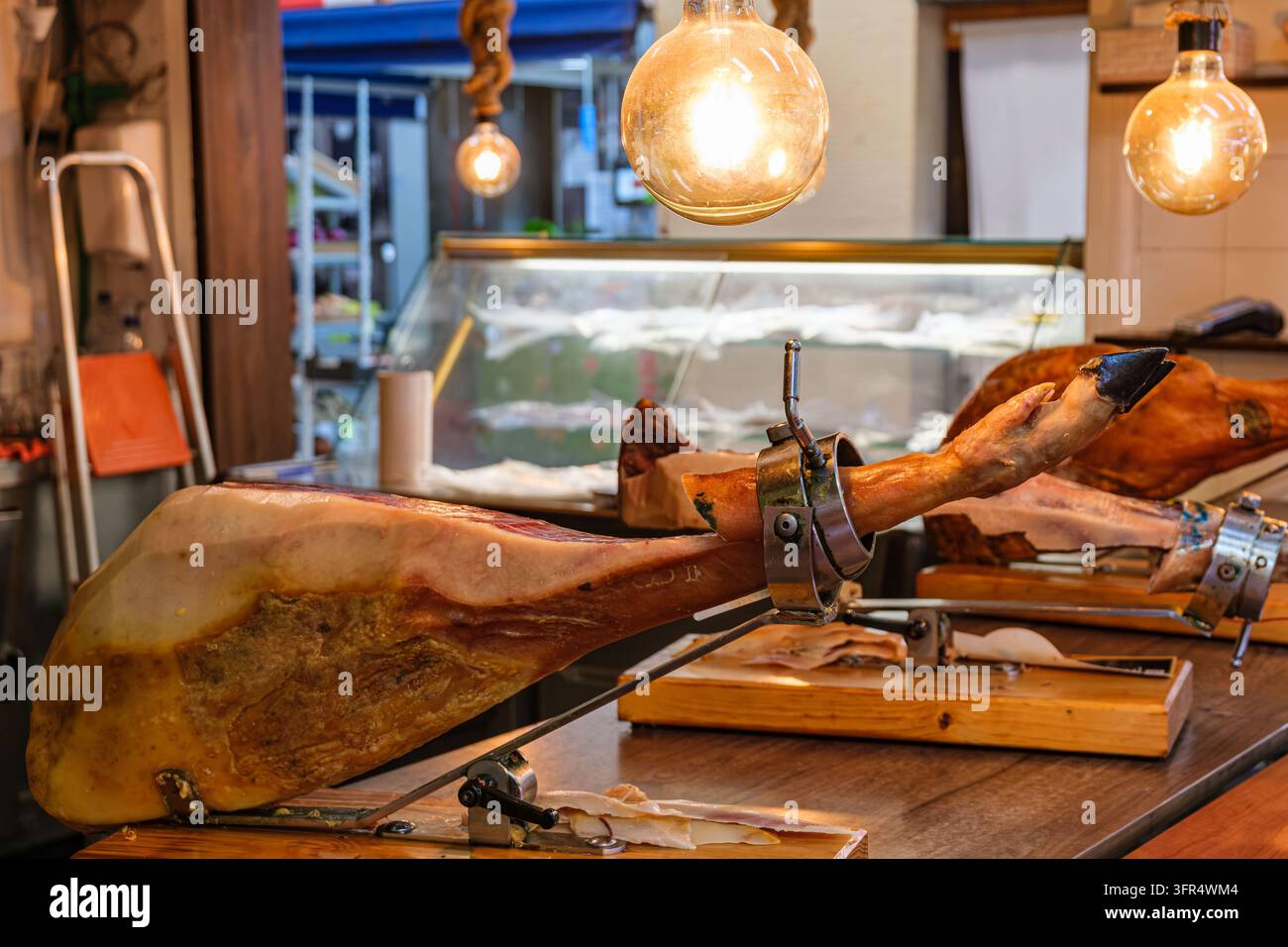 Ganzes Spanisches Schinkenbein in einem Holzschinkenständer (Jamonero) fixiert, beleuchtet von warmen Vintage-Edison-Glühbirnen an einer Delikatessen-Theke in einer Markthalle i Stockfoto