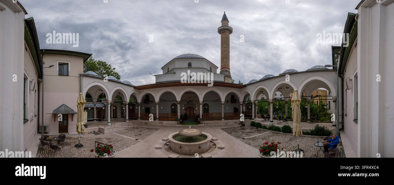 Panoramablick über den Hof der Kaiser-Moschee von Sarajevo, mit ihrer zentralen Kuppel und dem Minarett, die sich deutlich gegen einen bewölkten Himmel abheben. Stockfoto