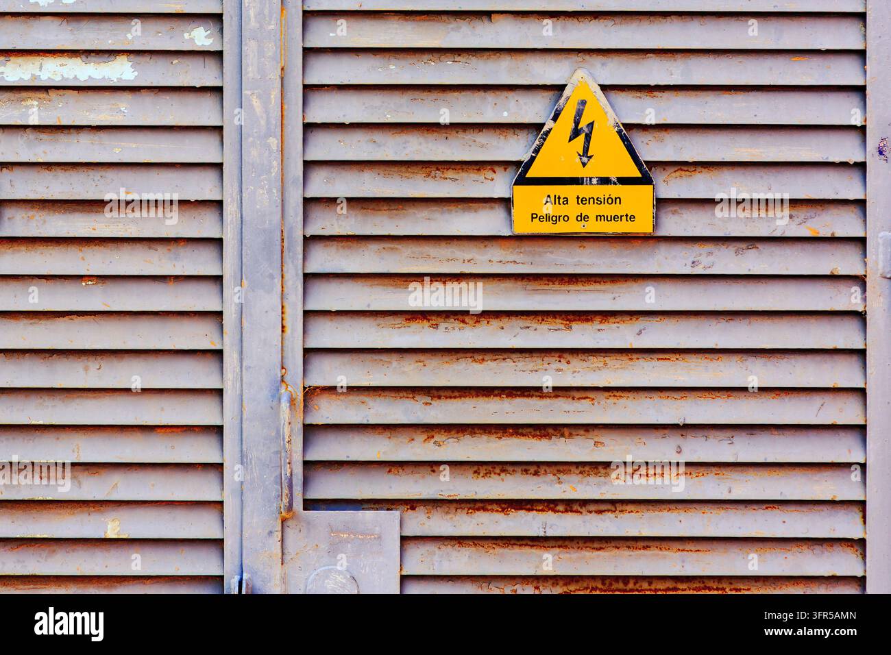 Zwei gelbe dreieckige Hochspannungs-Warnschilder sind auf einer verwitterten grauen metallenen Jalousien-Industrietür montiert. Jedes Schild zeigt einen schwarzen Blitz Stockfoto
