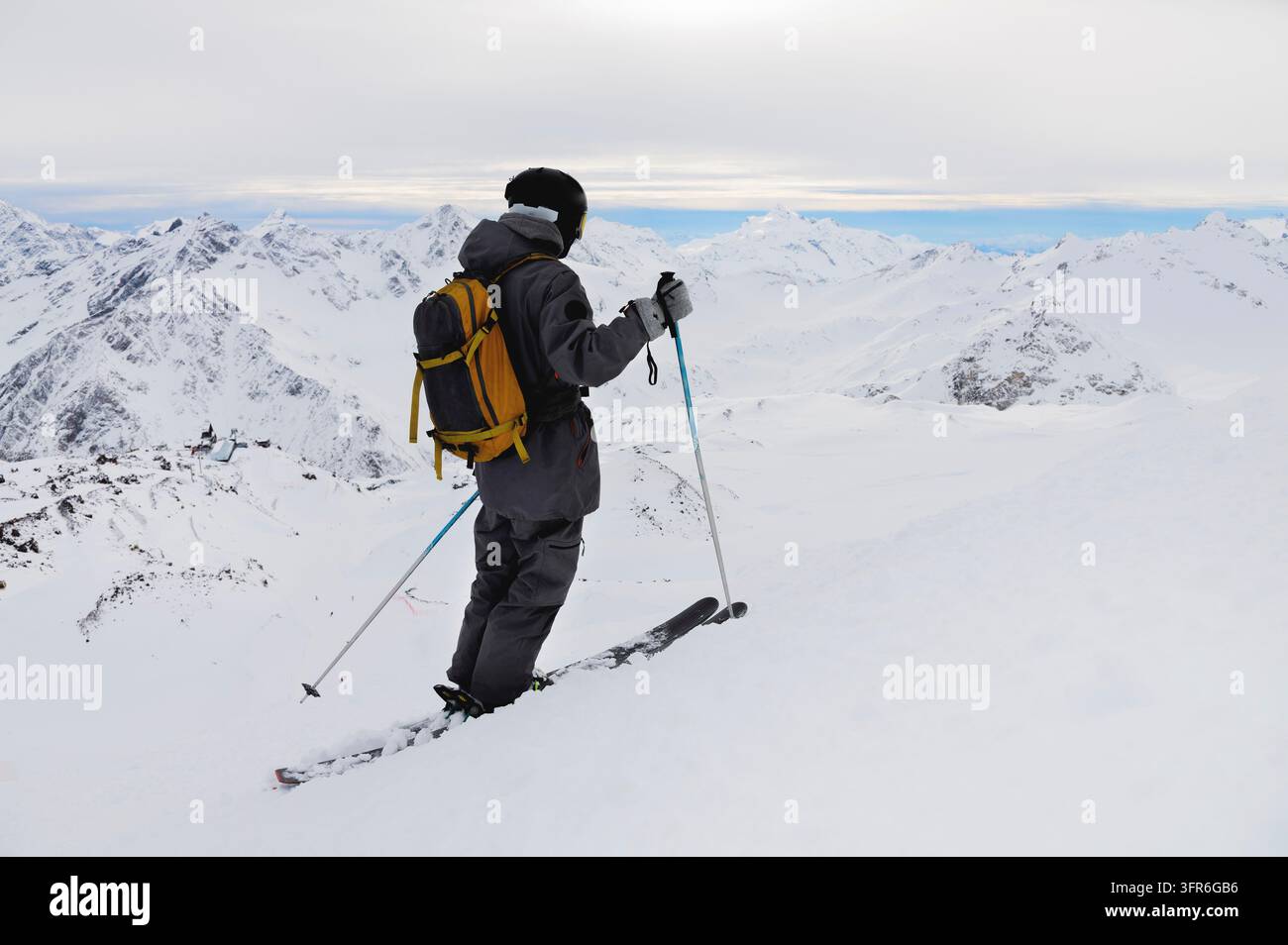 Seitenansicht eines sportlichen Skifahrers mit Helm und Skibrille sowie einem Rucksack, der auf Skiern steht, Skistöcke in der Hand hält, in weißem Schnee vor dem Himmel und eine Pause genießt Stockfoto