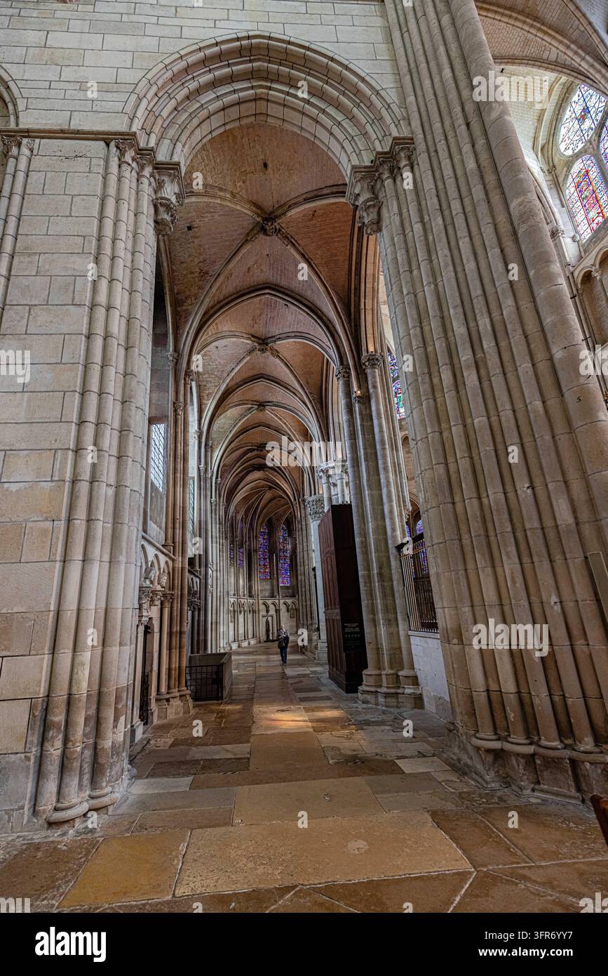 Ein Besucher geht entlang eines Kirchengangs in Auxerre, Frankreich, unter Rippengewölben und Steinbögen. Gruppenweise Säulen, geschnitzte Kapitelle und Buntglasfenster Stockfoto