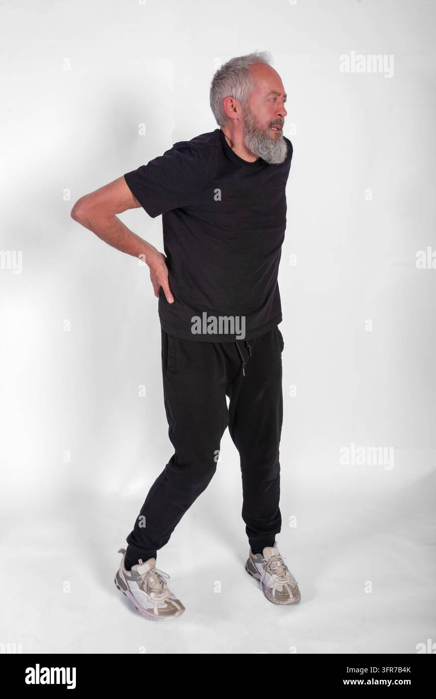 Eine Ganzkörper-Studioaufnahme eines reifen Mannes mit grauem Bart und schwarzer Sportkleidung, der vor Unbehagen grinst und sich mit beiden Händen den unteren Rücken hält Stockfoto
