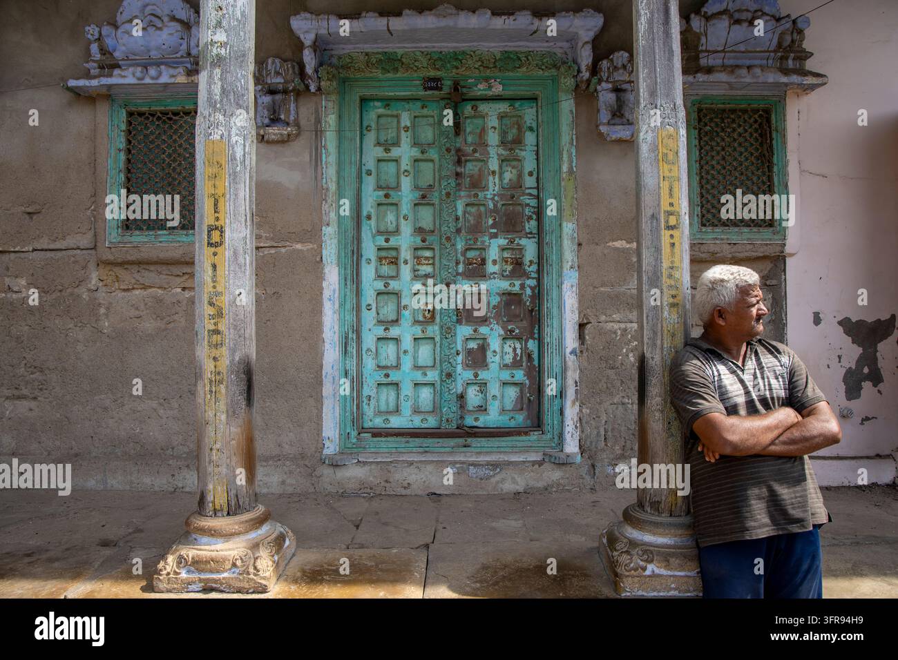 Ein älterer Mann steht mit verschränkten Armen neben einer Säule neben einer verwitterten türkisfarbenen geschnitzten Tür, Gujarat, Mandvi, Indien Stockfoto