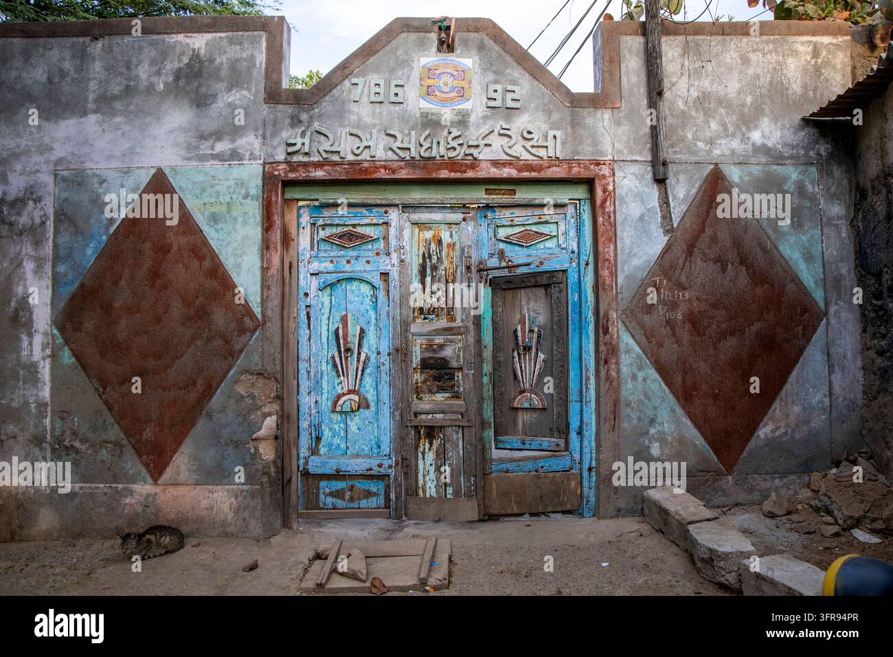Verwitterte blaue Holztür mit gujaratischer Inschrift und einer Katze, die in der Nähe auf dem Boden schläft, Gujarat, Mandvi, Indien Stockfoto
