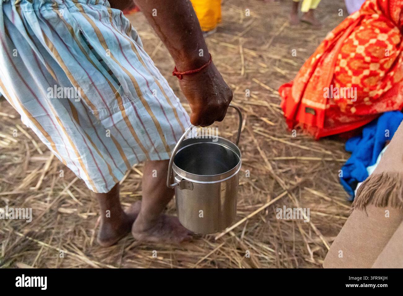 Nahaufnahme eines barfüßigen Mannes in einem gestreiften Lungi, der einen Stahlkübel auf strohbedecktem Boden bei der Maha Kumbh Mela in Uttar Pradesh, Prayagraj, Indien hält Stockfoto