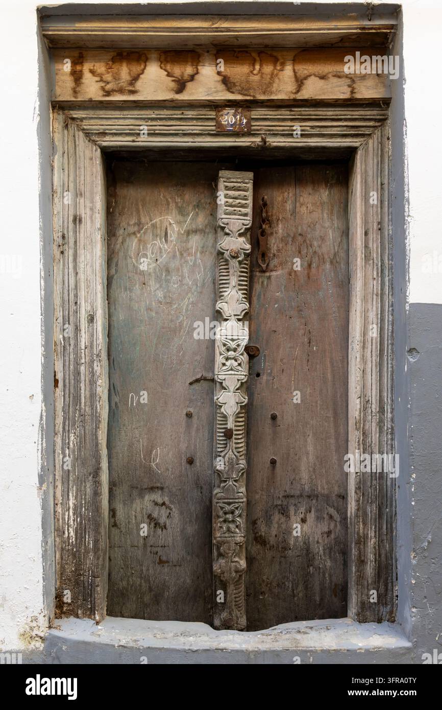 Verwitterte Holztür mit geschnitztem Mittelpfosten und altem Türsturz aus Holz, eingebettet in eine weiß getünchte und grau gestrichene Wand mit einem kleinen Namensschild, Sansibar Stockfoto