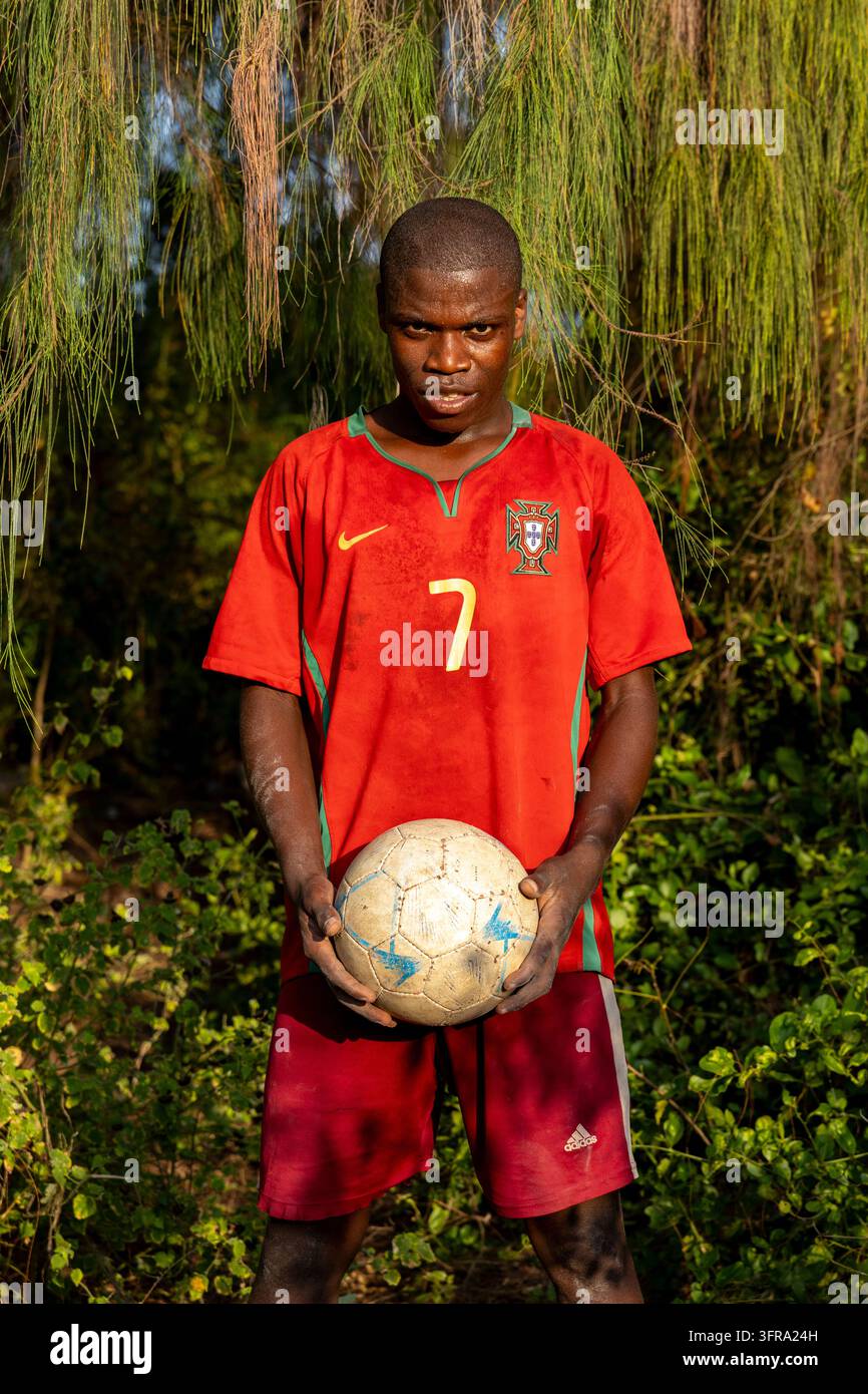 Junger Mann in einem roten Fußballtrikot von Portugal mit der Nummer sieben, der einen abgenutzten Fußball hält und unter hängenden Casuarina-Bäumen in warmem Licht in die Kamera schaut Stockfoto