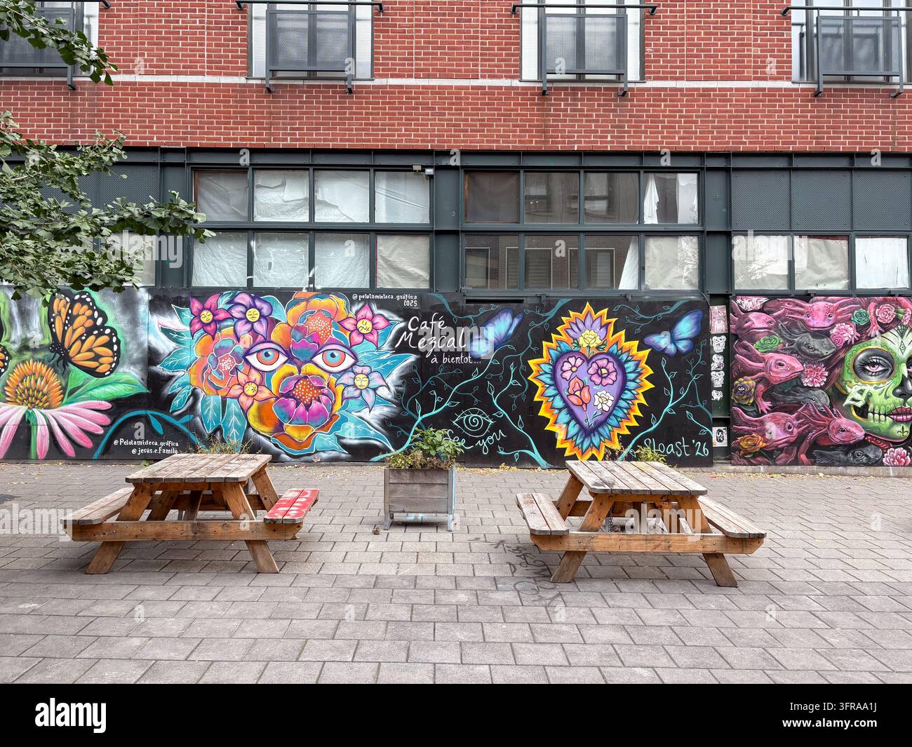 Farbenfroher Wandgemälde mit Café Mezcal-Kunstwerken, Schmetterlings- und Blumenmotiven, zusammen mit Holztischen auf einem Ziegelgebäudehof. Stockfoto