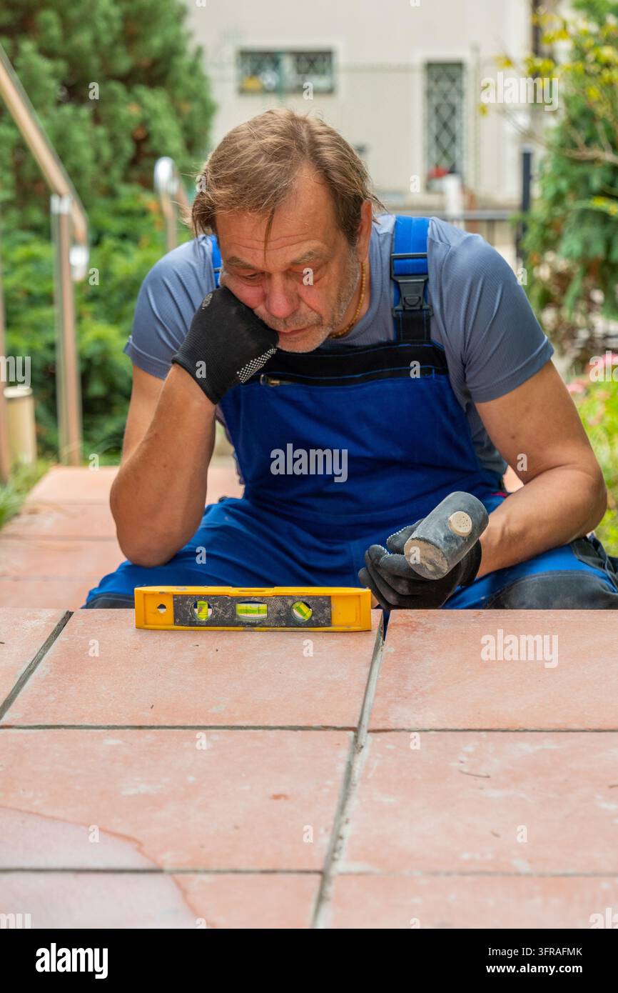 Ein müder Handwerker in blauen Latzhosen untersucht Außentreppenfliesen mit einem Hammer und einer Wasserwaage im Freien. Stockfoto