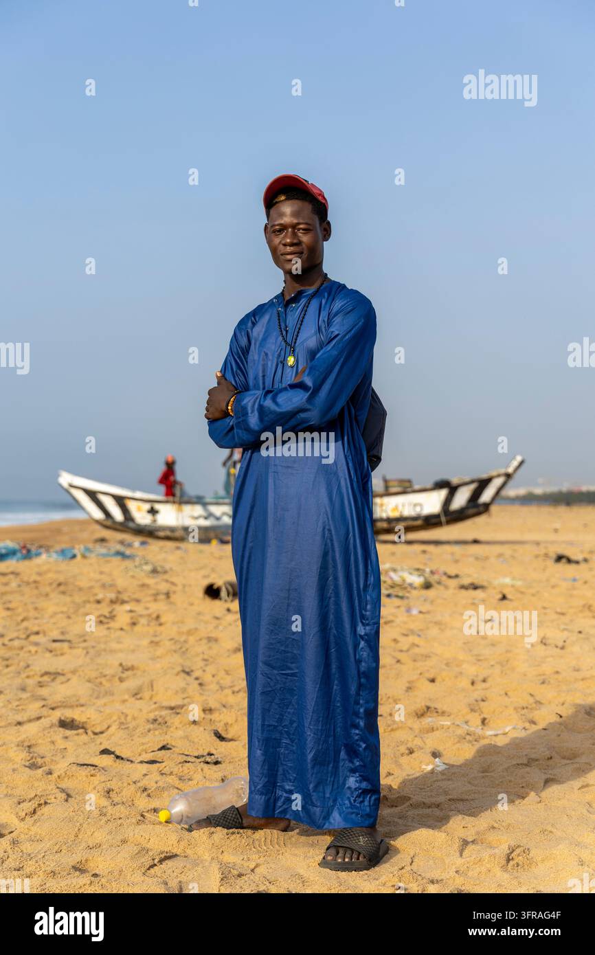 Porträt eines jungen Mannes in einem traditionellen blauen Gewand, der an einem Strand mit Fischerbooten im Hintergrund steht, Maritime Region, Lome, Togo Stockfoto