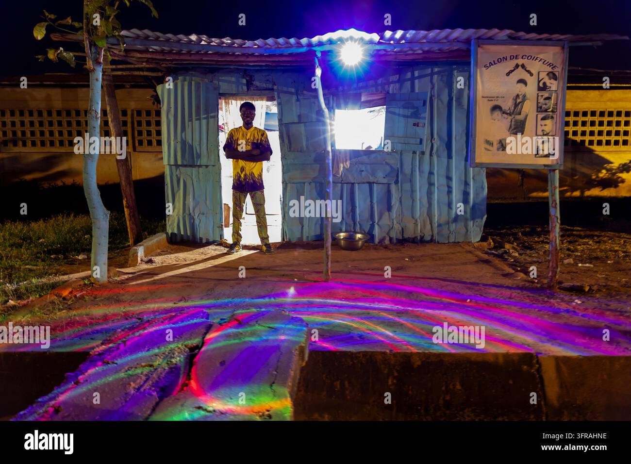 Mann steht in der Tür eines Straßenrandfriseursalons, der nachts von buntem Licht beleuchtet wird, Maritime Region, Togoville, Togo Stockfoto