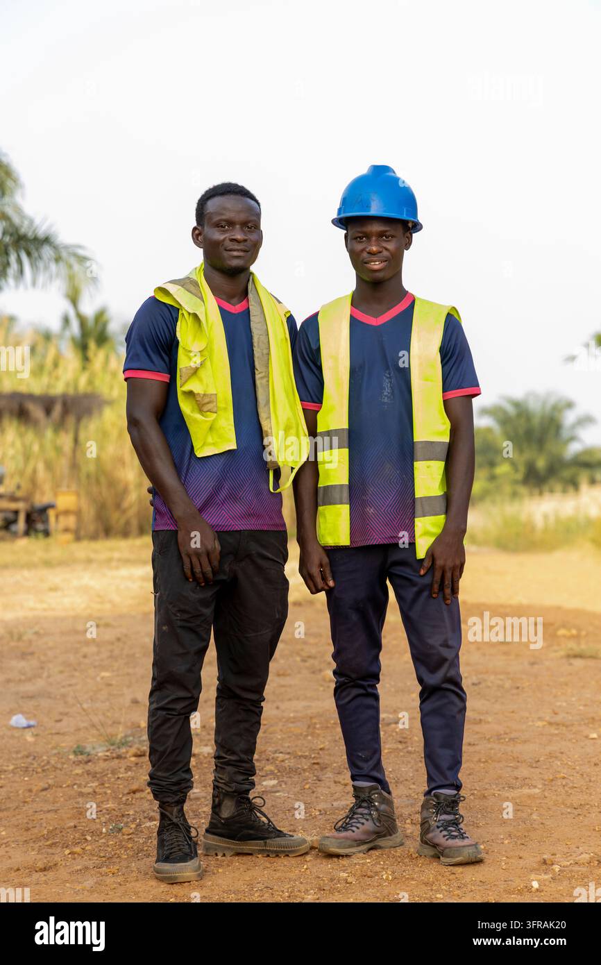 Zwei junge Bauarbeiter tragen gelbe Sicherheitswesten und einer trägt einen blauen Helm, während sie im Freien posieren, Plateaux-Region, Kpalime, Togo Stockfoto