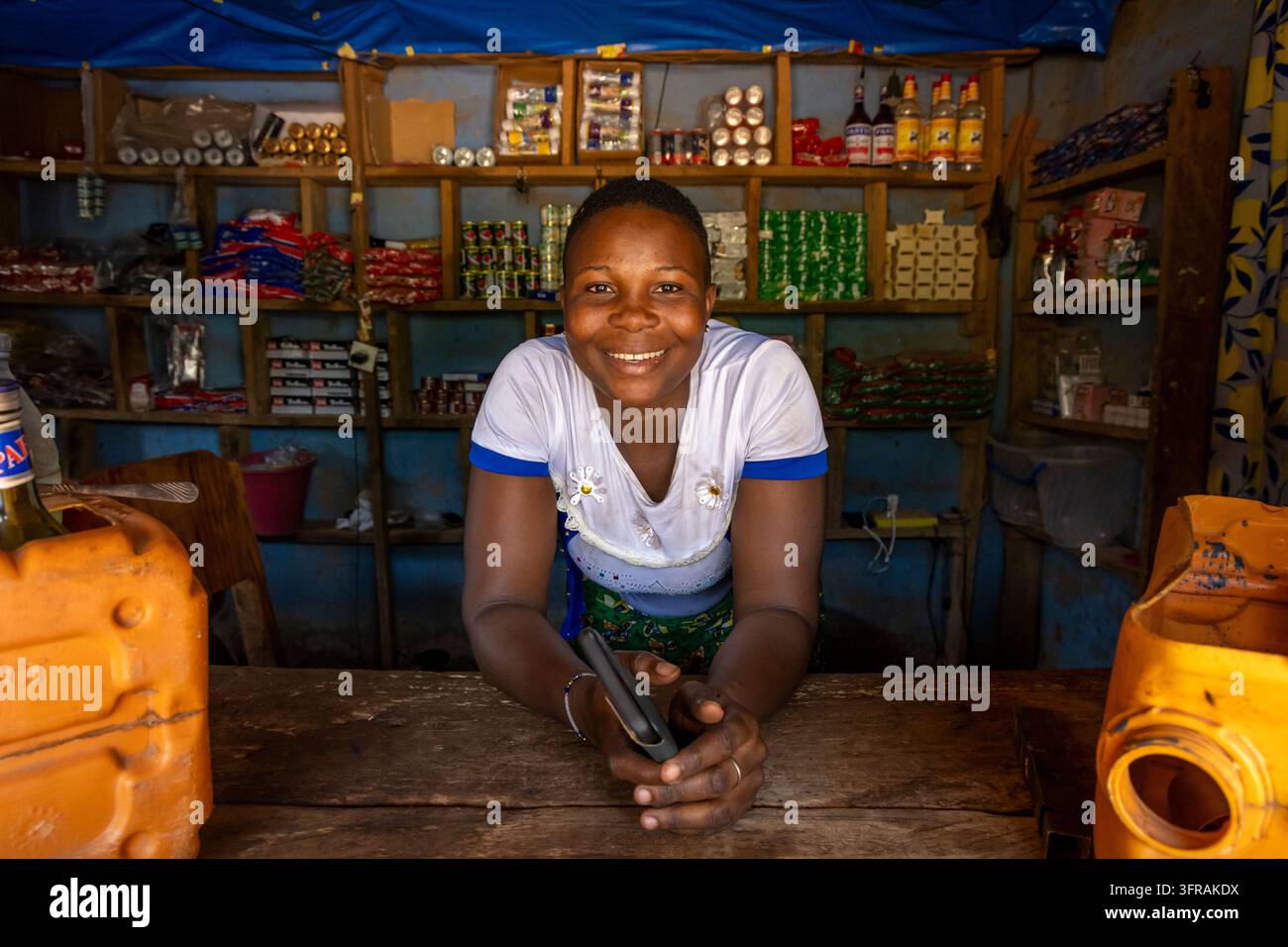 Frau lächelt, während sie sich auf eine Holztheke in einem kleinen Laden mit Waren in Koutammakou, Tamberma, Togo lehnt Stockfoto