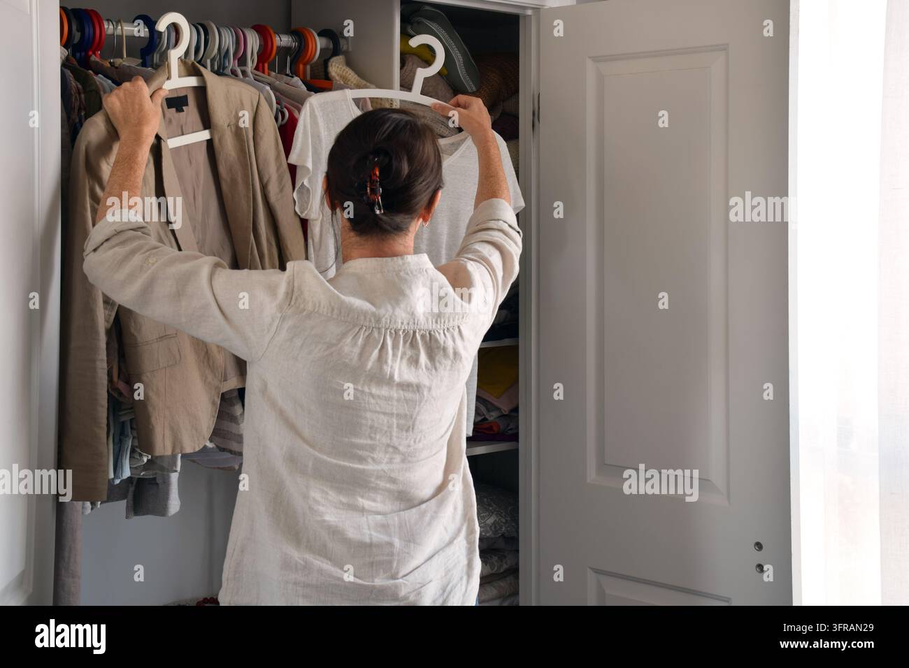 Rückansicht einer Frau, die einen Blazer und ein T-Shirt auf Kleiderbügeln hochhält und entscheidet, was sie aus ihrem Kleiderschrank anziehen soll Stockfoto