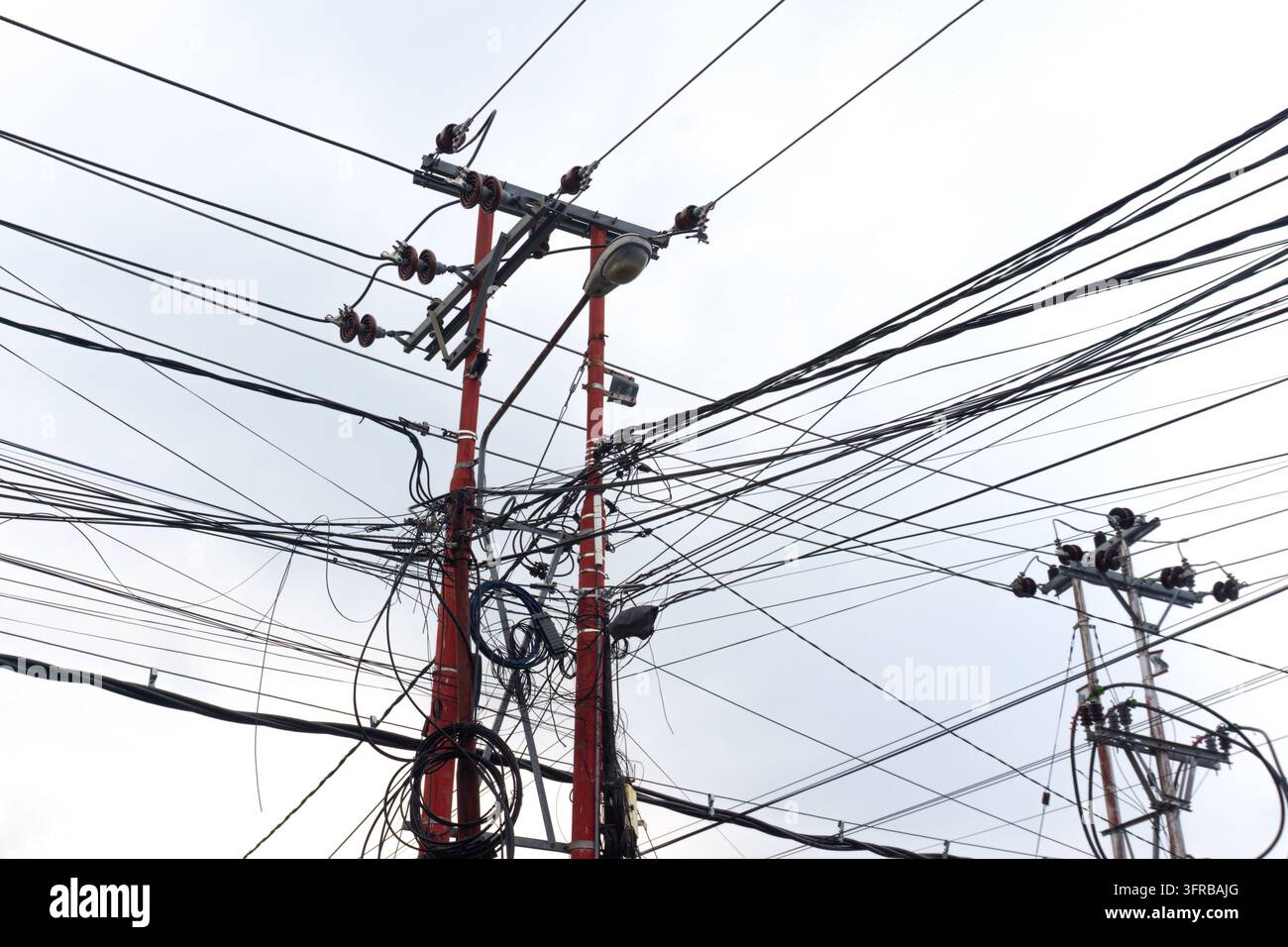Unordentliche Überkopfleitungen und Kabelsalat an Strommasten neben einer Straßenlaterne, bewölkter Himmel mit Kopierraum Stockfoto