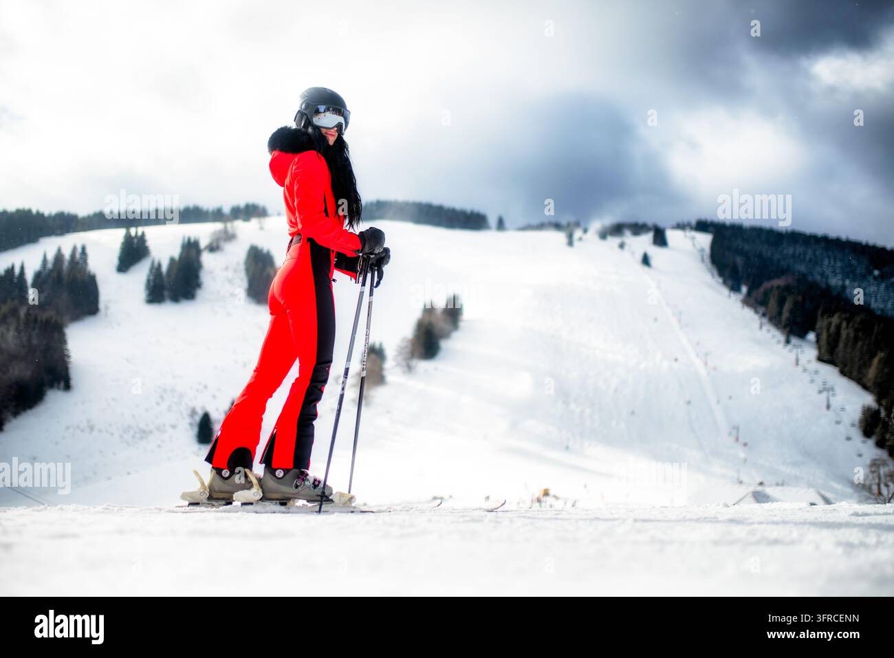 Ruzomberok, Slowakei - 4. Januar 2026: Skifahrerin in hellem rotem Skianzug mit Helm, Skibrille und Stöcken posiert auf einem schneebedeckten Hang am Malino Brdo Skigebiet Stockfoto
