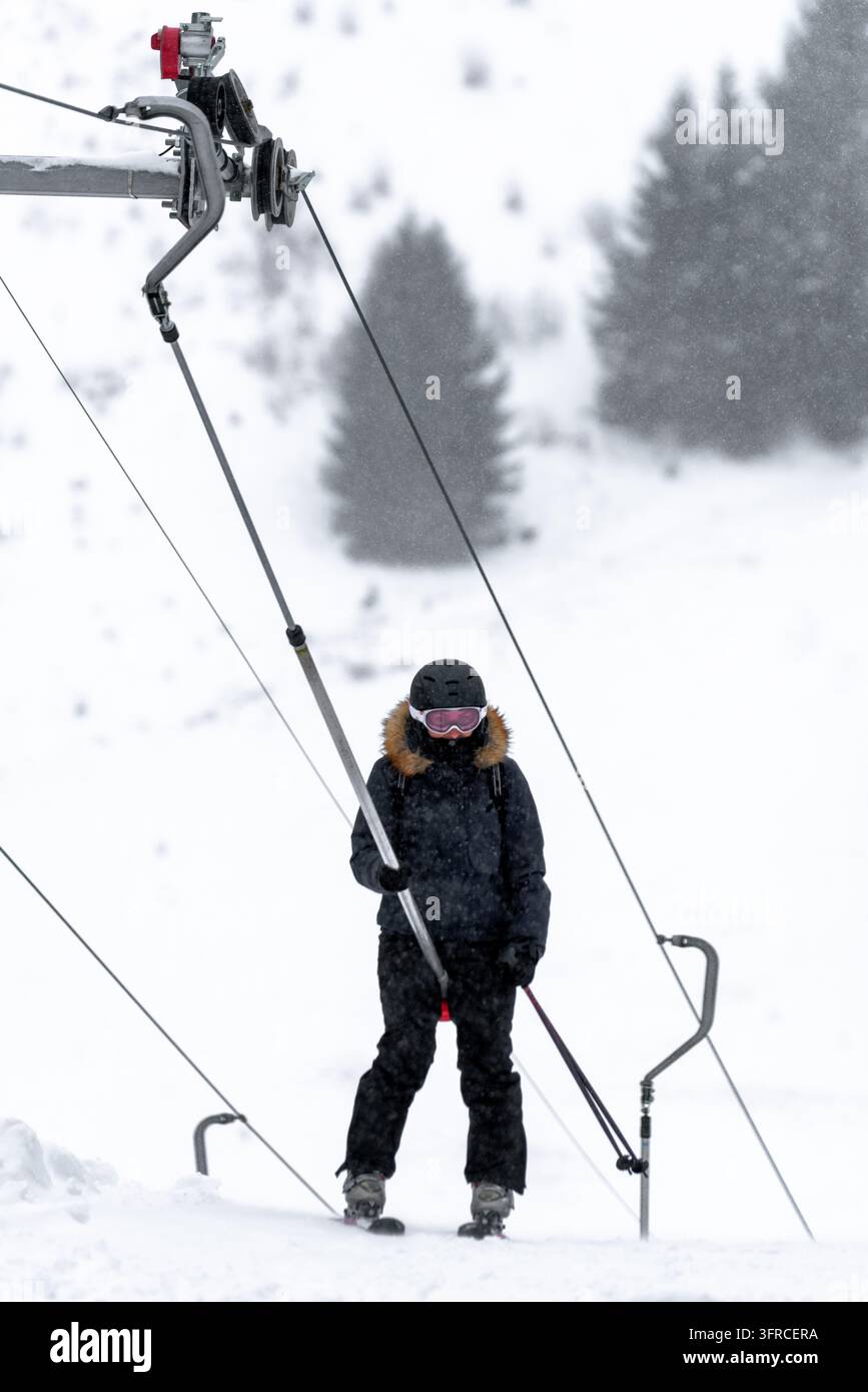Skifahrer in schwarzer Winterkleidung, Helm und rosa Skibrille fährt einen oberflächlichen Skilift während des Schneefalls, mit schneebedeckten Berghängen und verschwommenem Hintergrund Stockfoto