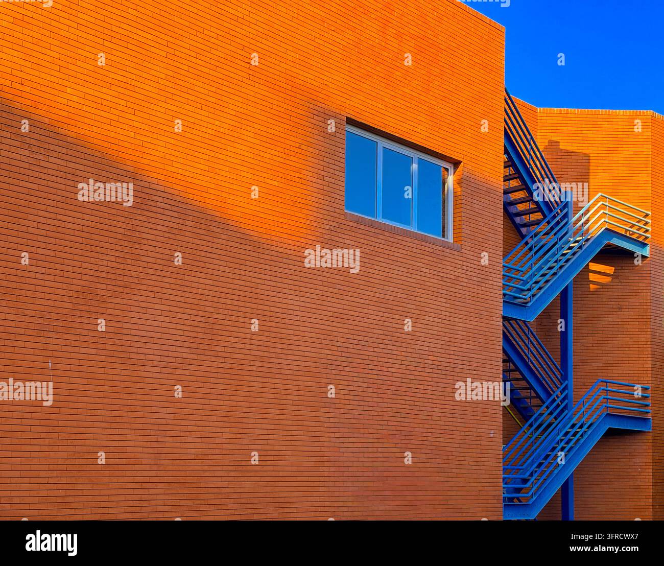 ORANGE FARBENE STEINFASSADE mit blauen Stahl-Feuerwehrtreppen, modernistische geometrische Gestaltung, starke Schatten und blauer Himmel Stockfoto