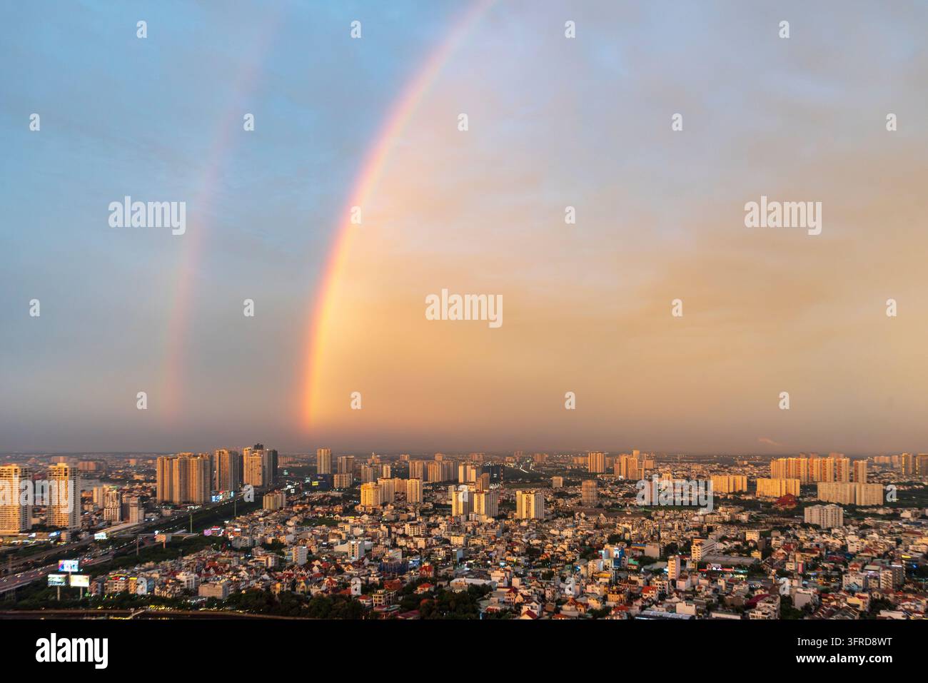 Dramatischer Sonnenuntergangshimmel mit einem lebhaften doppelten Regenbogen, der über den städtischen Gebäuden aufsteigt. Stockfoto