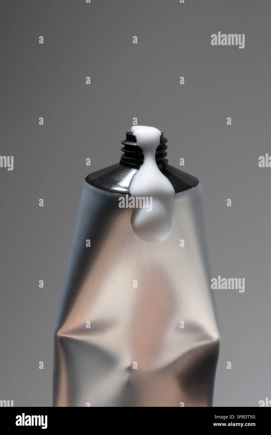 Ein schlankes, silbernes Metallröhrchen für Sahne, das ausgepresst wird, um die Produktanwendung auf einem grauen Hintergrund zu zeigen. Saubere, moderne und minimalistische Produktfotografie, ideal für Co Stockfoto