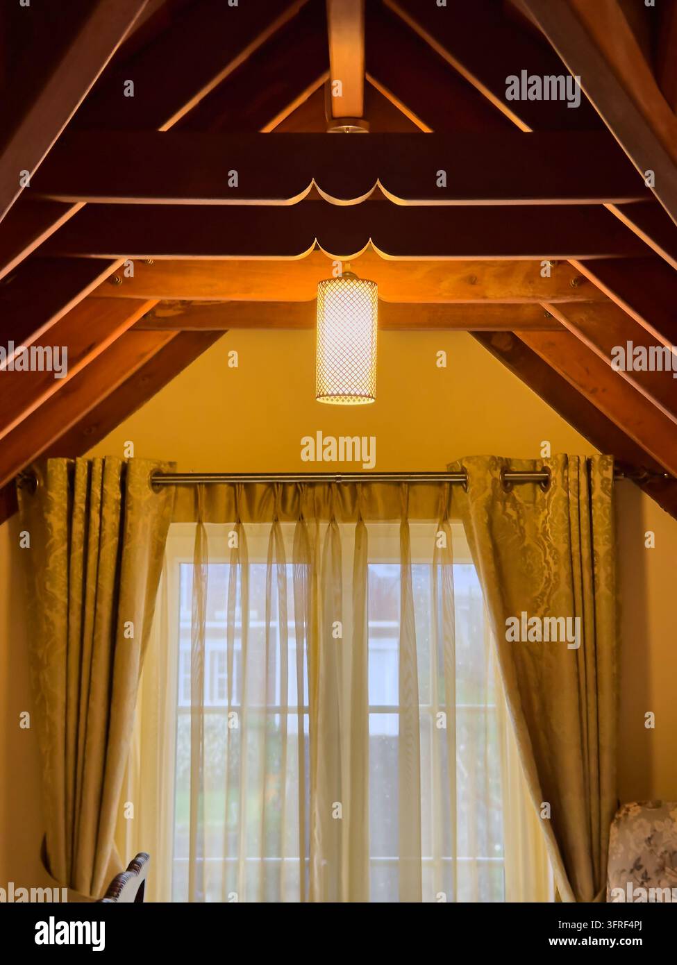 warmes Holzhaus-Interieur mit A-Frame-Decke, hängender zylindrischer Lampe und Fenster mit durchsichtigen Vorhängen, gemütliches rustikales Wohnkonzept Stockfoto