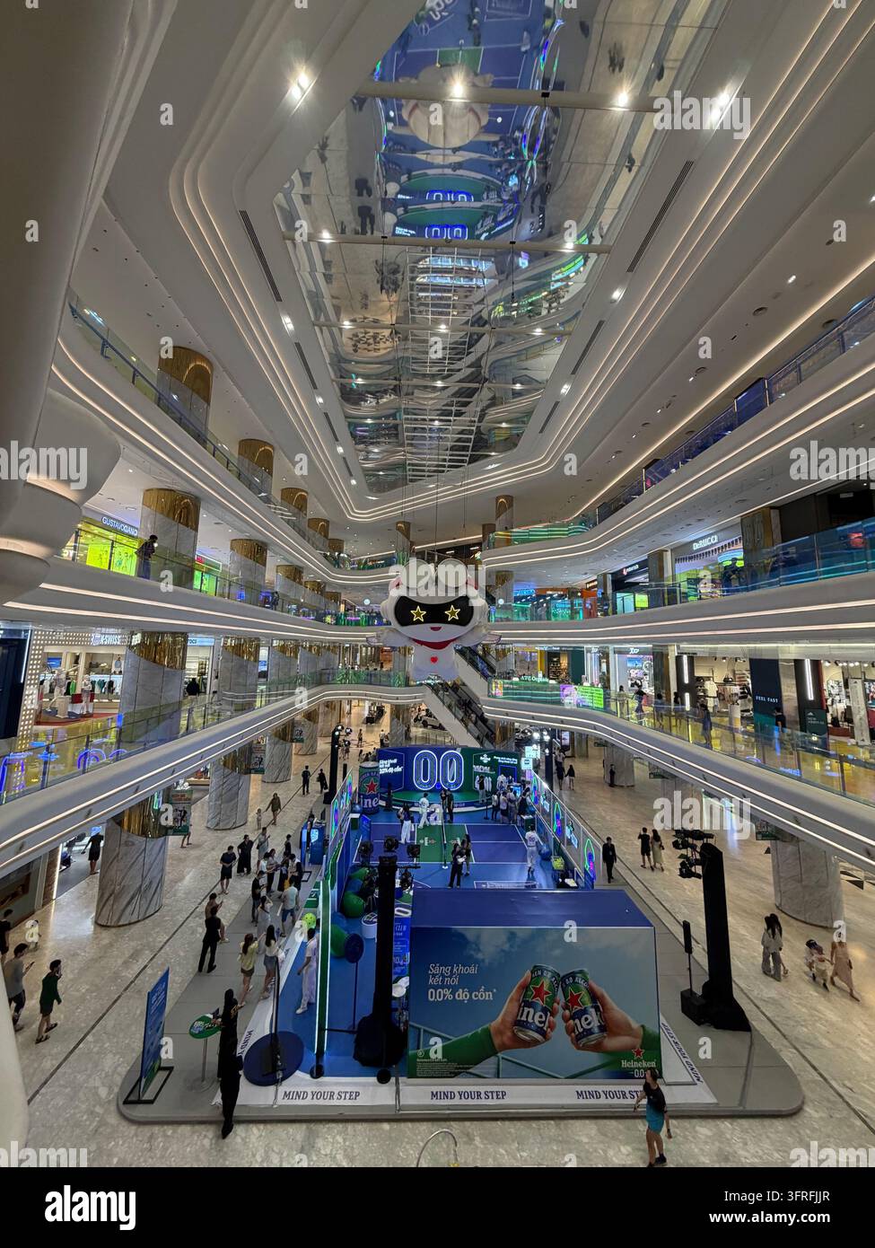 Eine weitläufige Innenansicht zeigt Einkäufer, geschwungene Balkone, Rolltreppen, Schaufenster, eine Werbeinstallation und Reflexionen an einer verspiegelten Decke in einem modernen mehrstöckigen Einkaufszentrum. Stockfoto