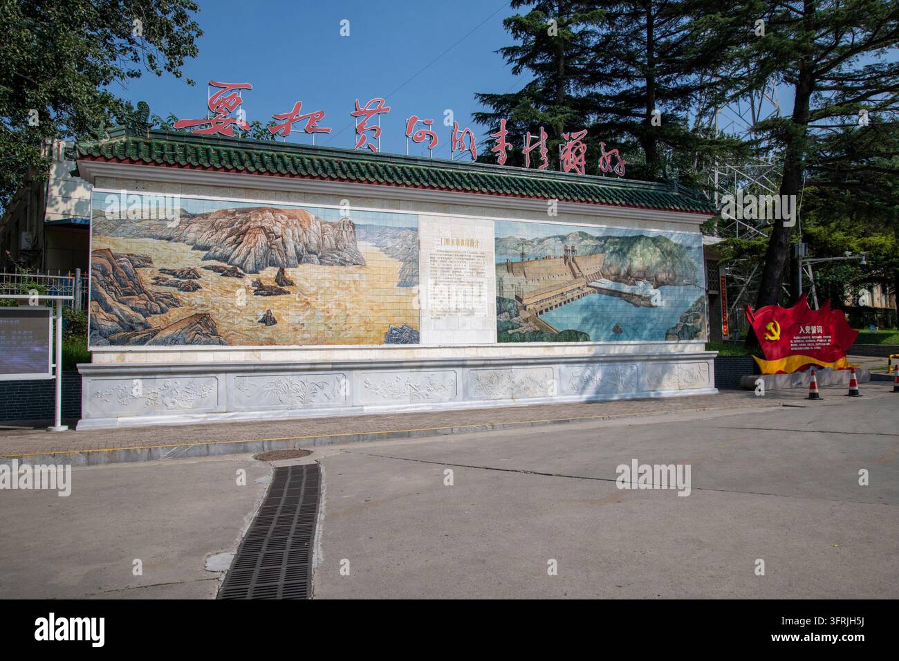 Eine dekorative Wand mit großen bildlichen Reliefpaneelen und chinesischer Schrift zwischen Bäumen in Sanmenxia, Henan, China. Stockfoto
