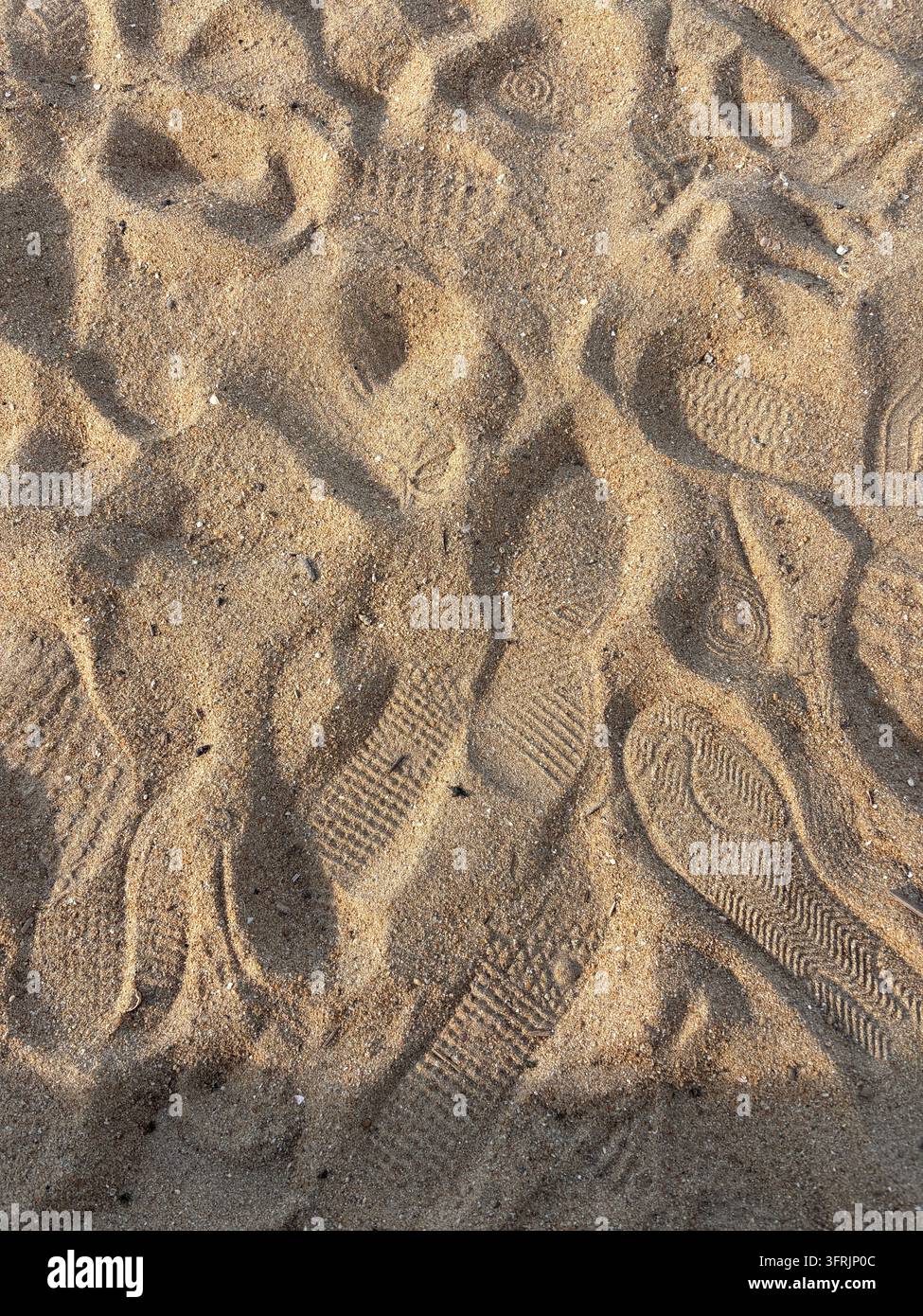 Hohe Winkelaufnahme von zahlreichen verschiedenen Fußabdrücken und Schuhsohlenmustern, die sich im goldenen Strandsand am Momochi-Strand in Fukuoka abzeichnen. - Smartphone-aufgenommenes Stockfoto