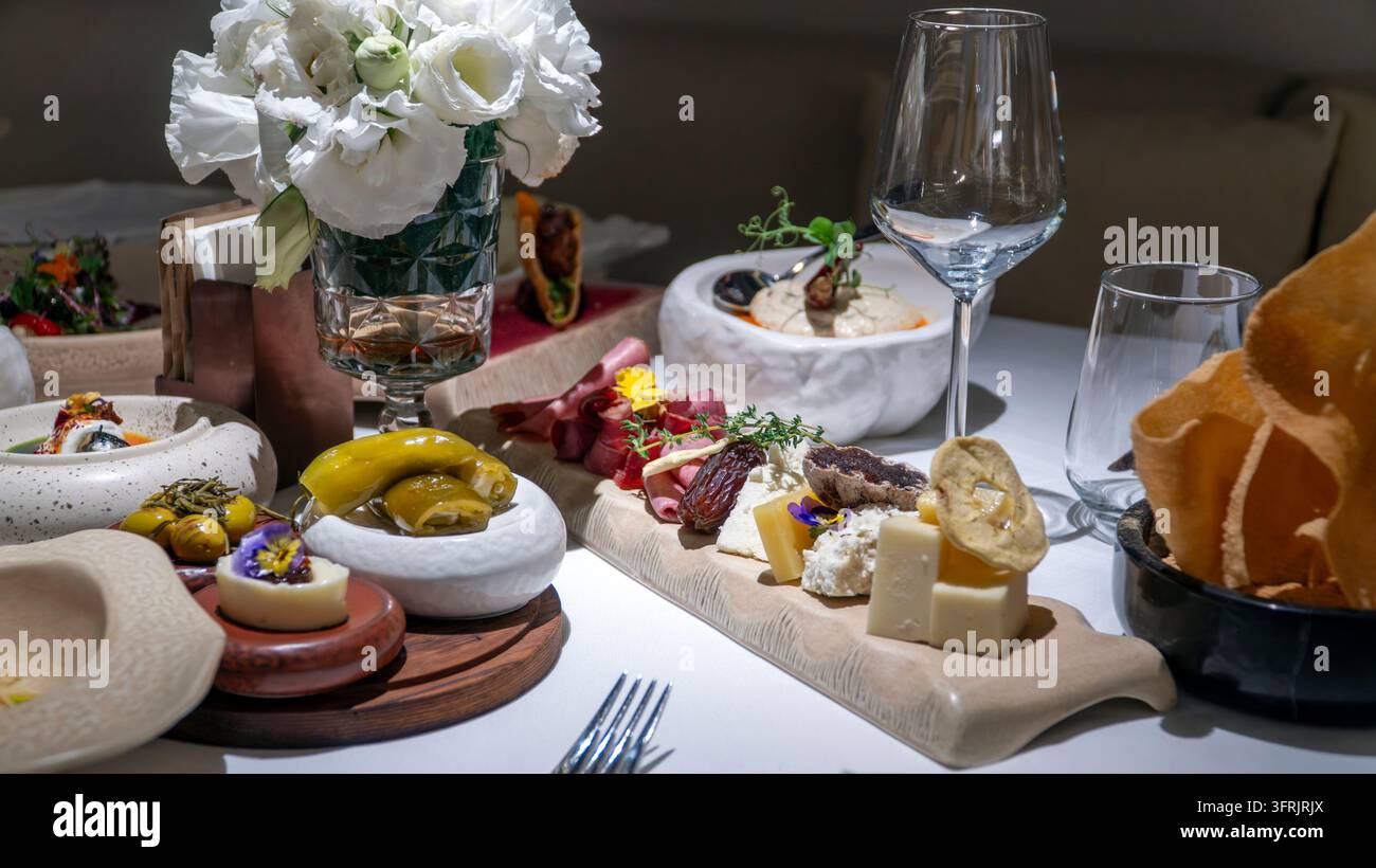 Schneidebrett mit Käsewürfeln, gepökeltem Fleisch, Datteln und essbaren Blumen, mit Oliven, weißen Blumen und Weingläsern auf einem Restauranttisch in Turke Stockfoto