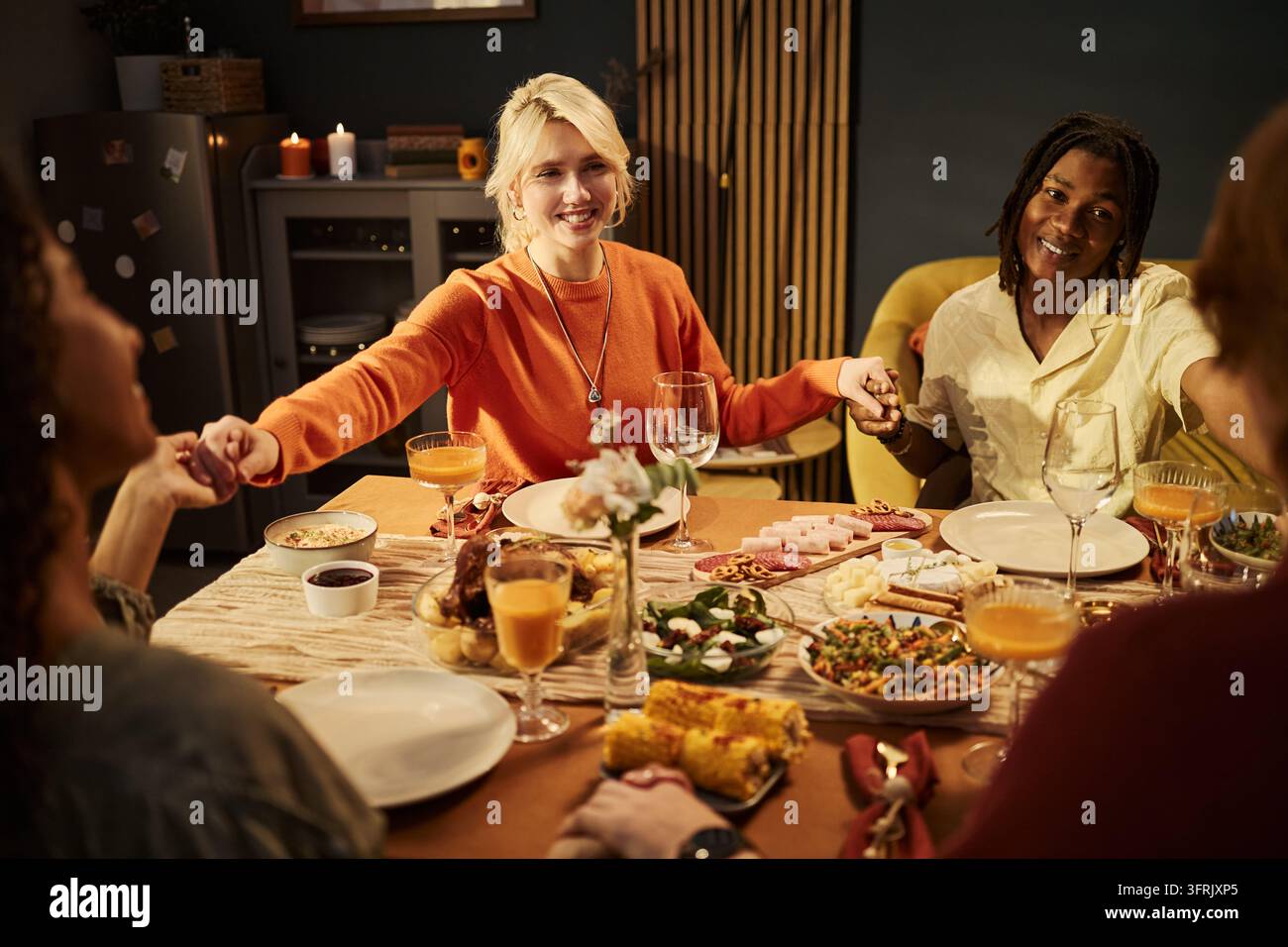 Verschiedene Freunde, die sich an einem Esstisch an den Händen halten und beten Stockfoto