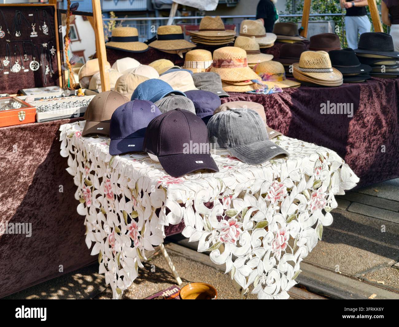 Bad Wildbad, Deutschland. Reihe von Baseballmützen und Strohhüten, die auf einem mit floraler Spitze bedeckten Markttisch im Freien angeordnet sind, sonnige Szene, die dunkles n zeigt Stockfoto