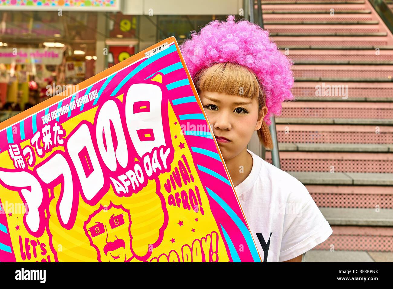 Junge japanische Frau mit einer lockigen rosa Perücke lugt hinter einem hellen Werbeposter vor einer rosa Treppe und einem Ladenfront in Japan hervor. Stockfoto