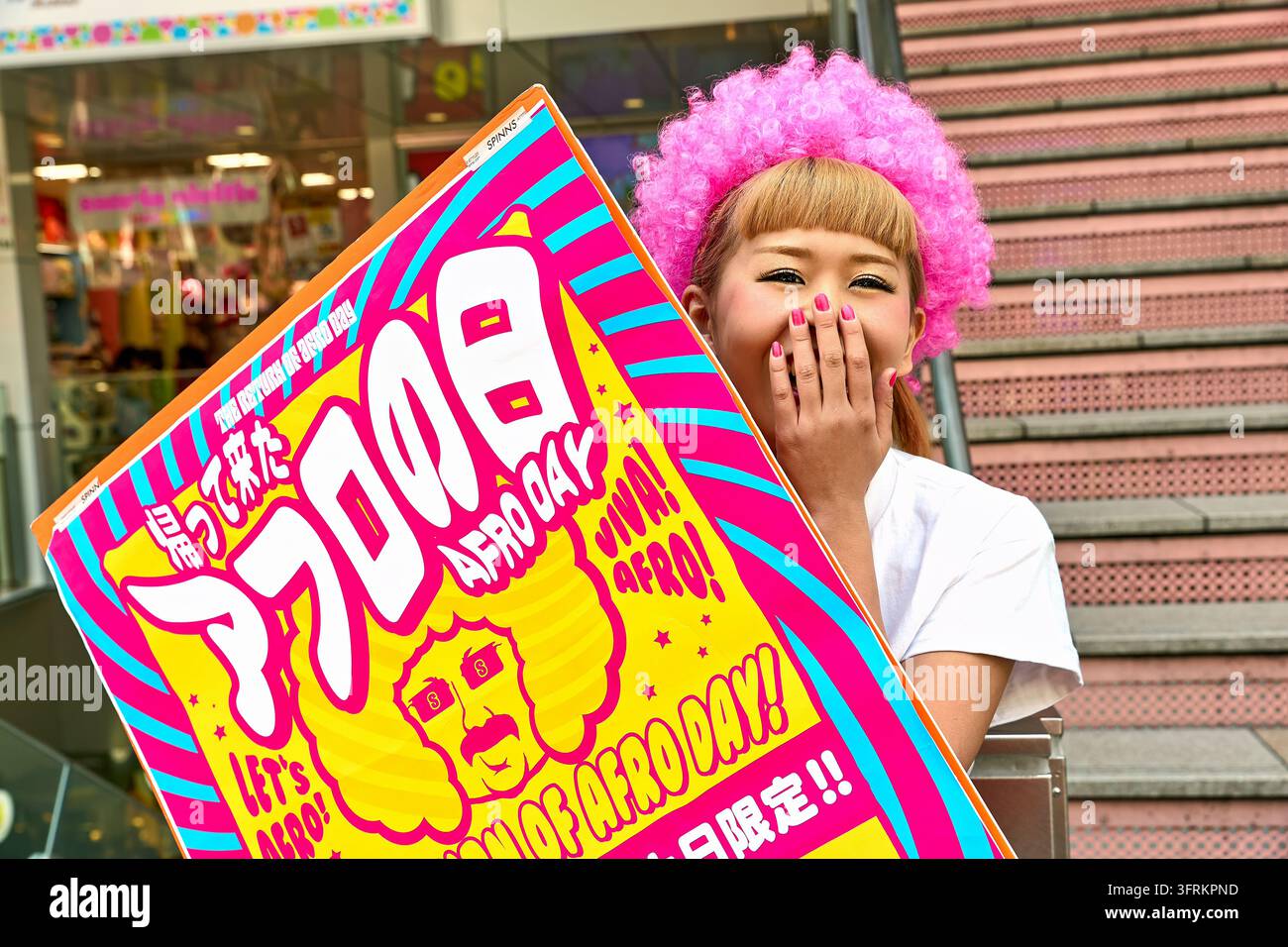 Junge Frau verbirgt ein Lachen hinter ihrer Hand, während sie ein gelbes und magentafarbenes Werbeschild für ein eintägiges Afro-Day-Event in Japan hält Stockfoto