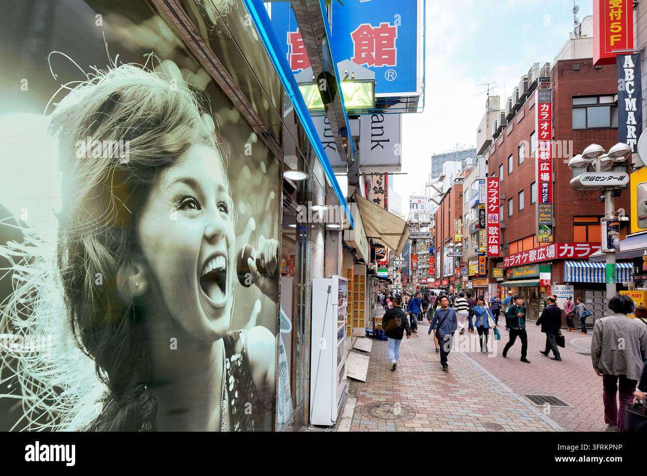 Fußgänger passieren Karaoke-Schilder und ein riesiges Plakat einer singenden Frau an einem Frühlingsnachmittag in Shibuya, einem Jugendkultur-Zentrum der japanischen Hauptstadt. Stockfoto