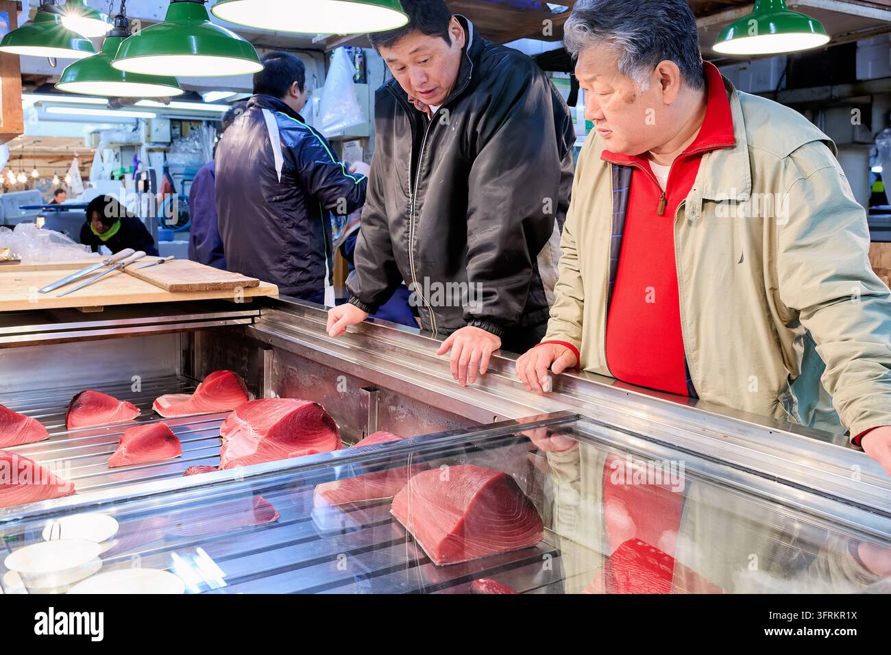 Zwei Kunden lehnen sich über eine gekühlte Glastheke mit rohen Thunfischblöcken, während Arbeiter Meeresfrüchte im Tokyo Fish Market zubereiten Stockfoto