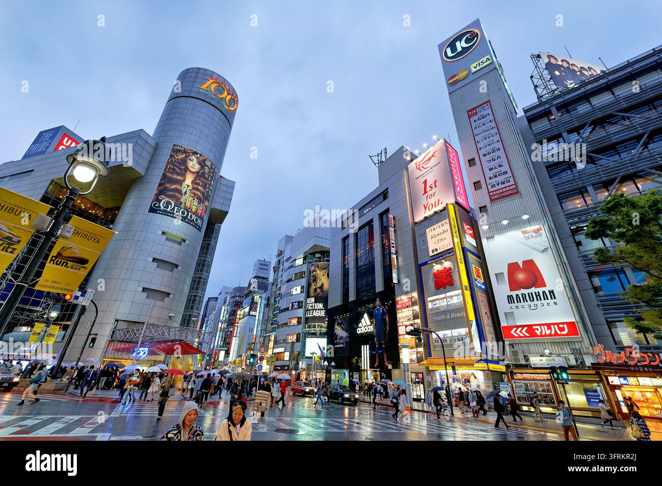Fußgänger mit Regenschirmen überqueren eine regennasse Kreuzung unter beleuchteten Werbetafeln im Einkaufsviertel Shibuya an einem Frühlingsabend. Stockfoto