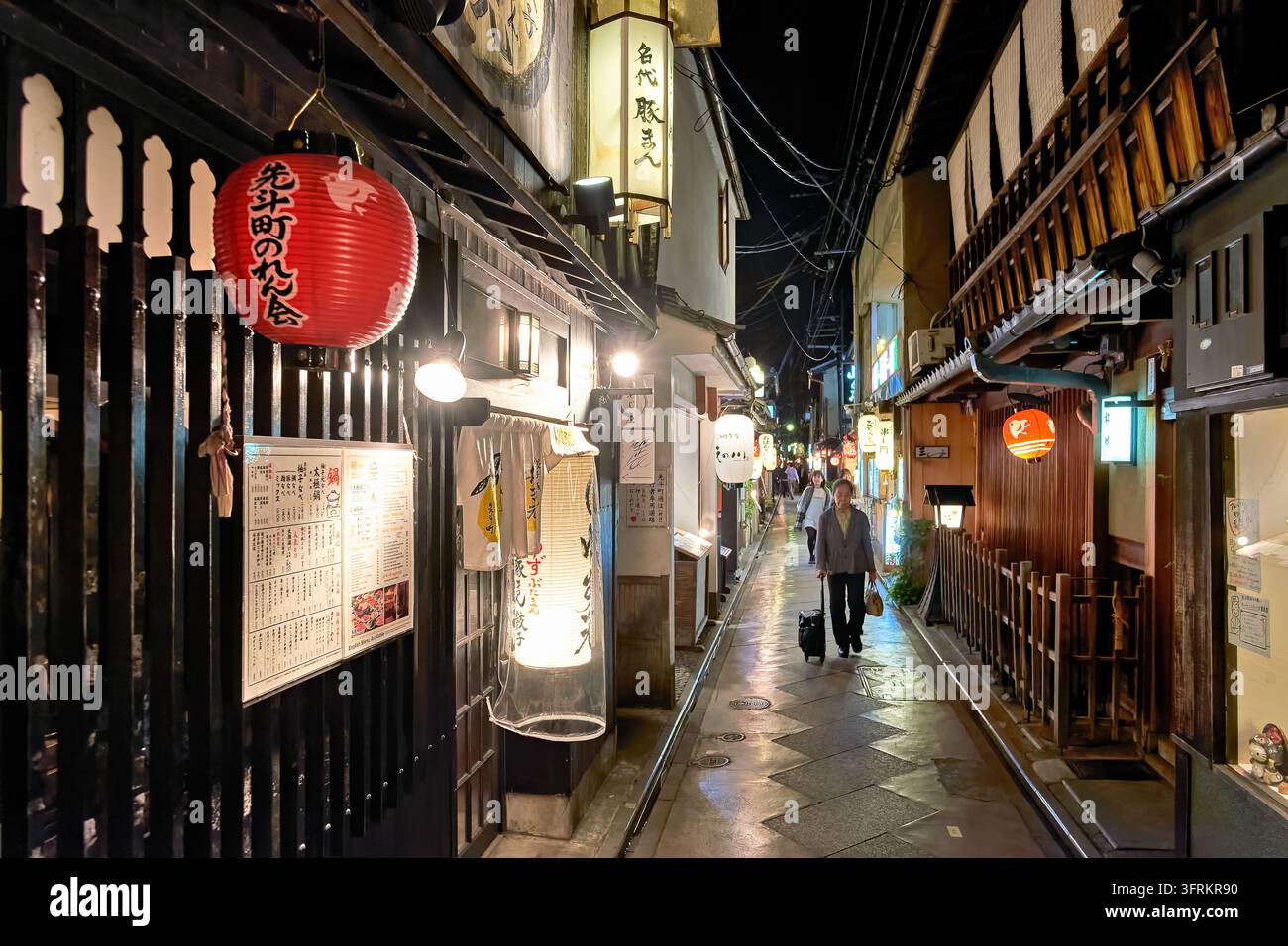 Frau mit einem Rollkoffer geht eine regennasse Gasse entlang, die von traditionellen Holzrestaurants im Pontocho-Essensviertel in Kyoto, Japan, gesäumt ist Stockfoto