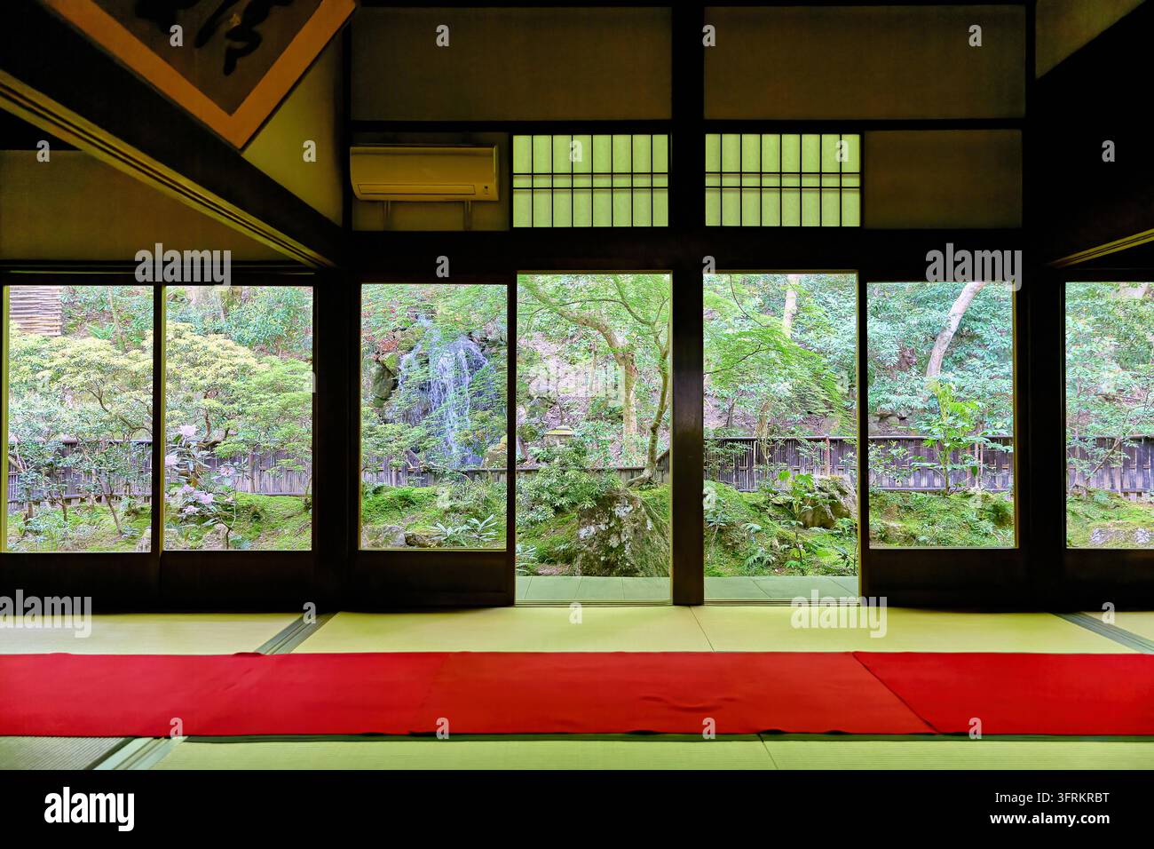 Rote Filzteppiche auf Tatami in einem traditionellen Tempelintern, mit offenen Fenstern, die auf einen Frühlingsgarten mit Steinlaterne und Ahornbäumen blicken. Stockfoto