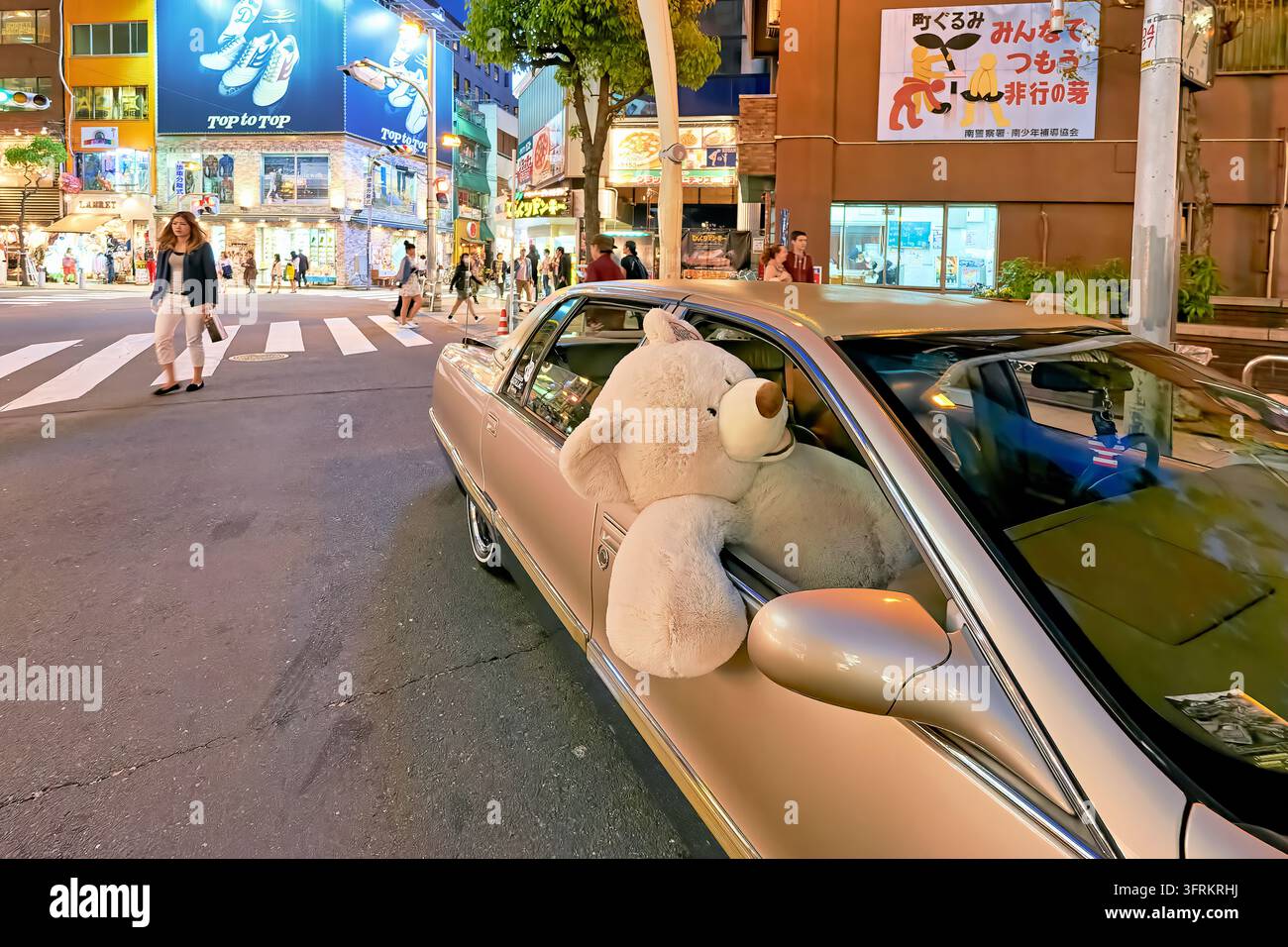 Übergroßer Plüschbär lehnt sich aus einem geparkten Sedan an einem Zebrastreifen im Minami-Bezirk von Osaka, Japan, an einem Frühlingsabend, während Passanten vorbeigehen Stockfoto