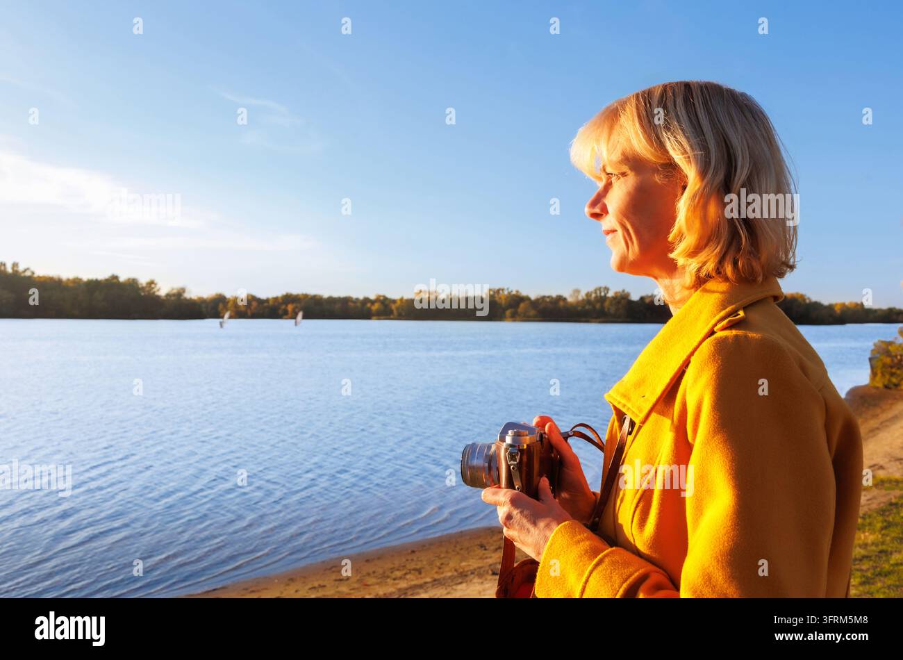 Reife Frau hält eine Vintage-Filmkamera und blickt friedlich über den See im warmen Abendlicht. Konzept des Digital Detox, eine Pause vom Informat Stockfoto