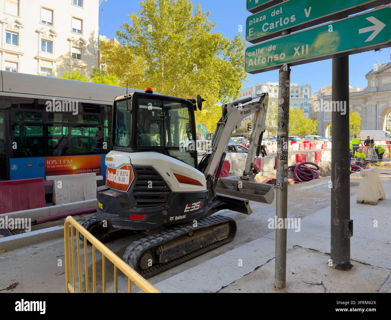 MADRID, SPANIEN - 09. OKTOBER 2026 Bobcat E35-Bagger steht bei Straßenarbeiten in Madrid neben einem Bus, mit Barrieren, Rohren und Arbeitern. Grüne Schilder zeigen Pla Stockfoto