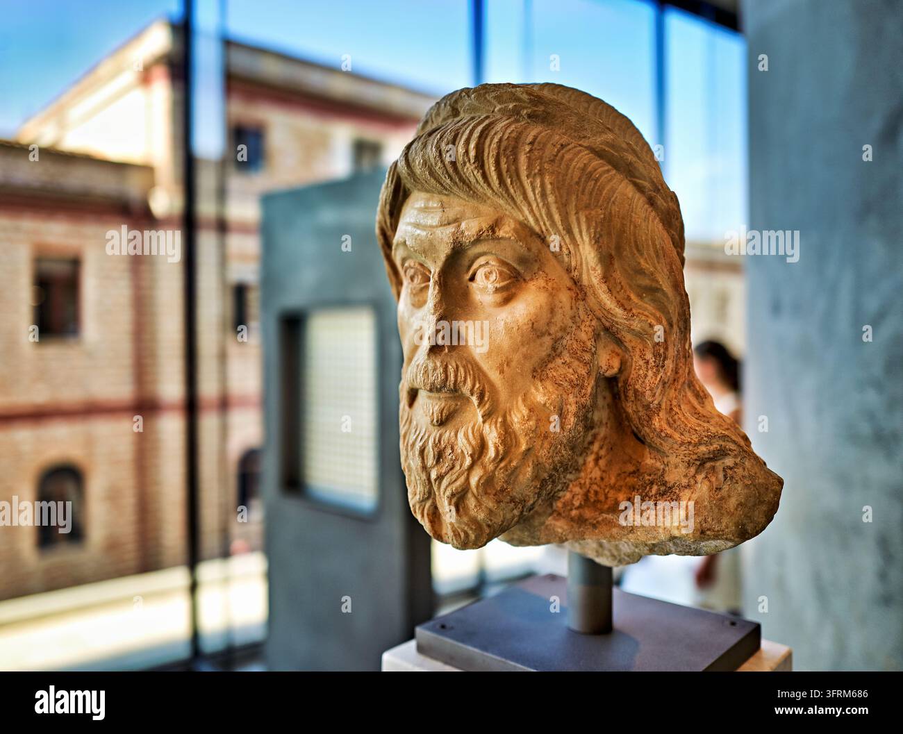 Antikes geschnitztes Porträt mit langen Haaren und Bart auf einer Stahlmontage neben einer Glasfassade, mit Blick auf ein Backsteingebäude in Griechenland Stockfoto